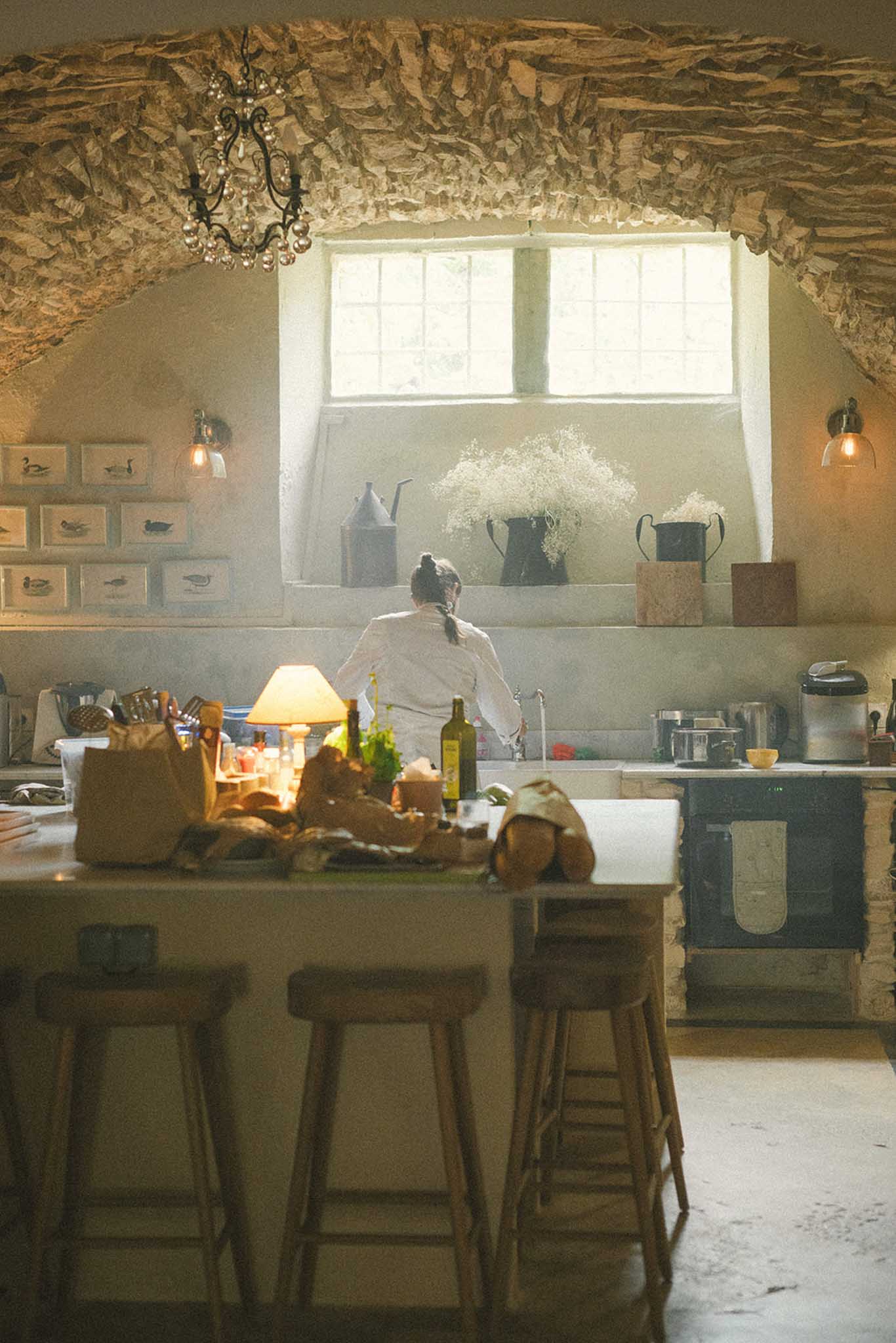 Bride in cream robe seen from behind at counter in rustic stone kitchen with wrought-iron chandelier, wine bottles, and white flowers