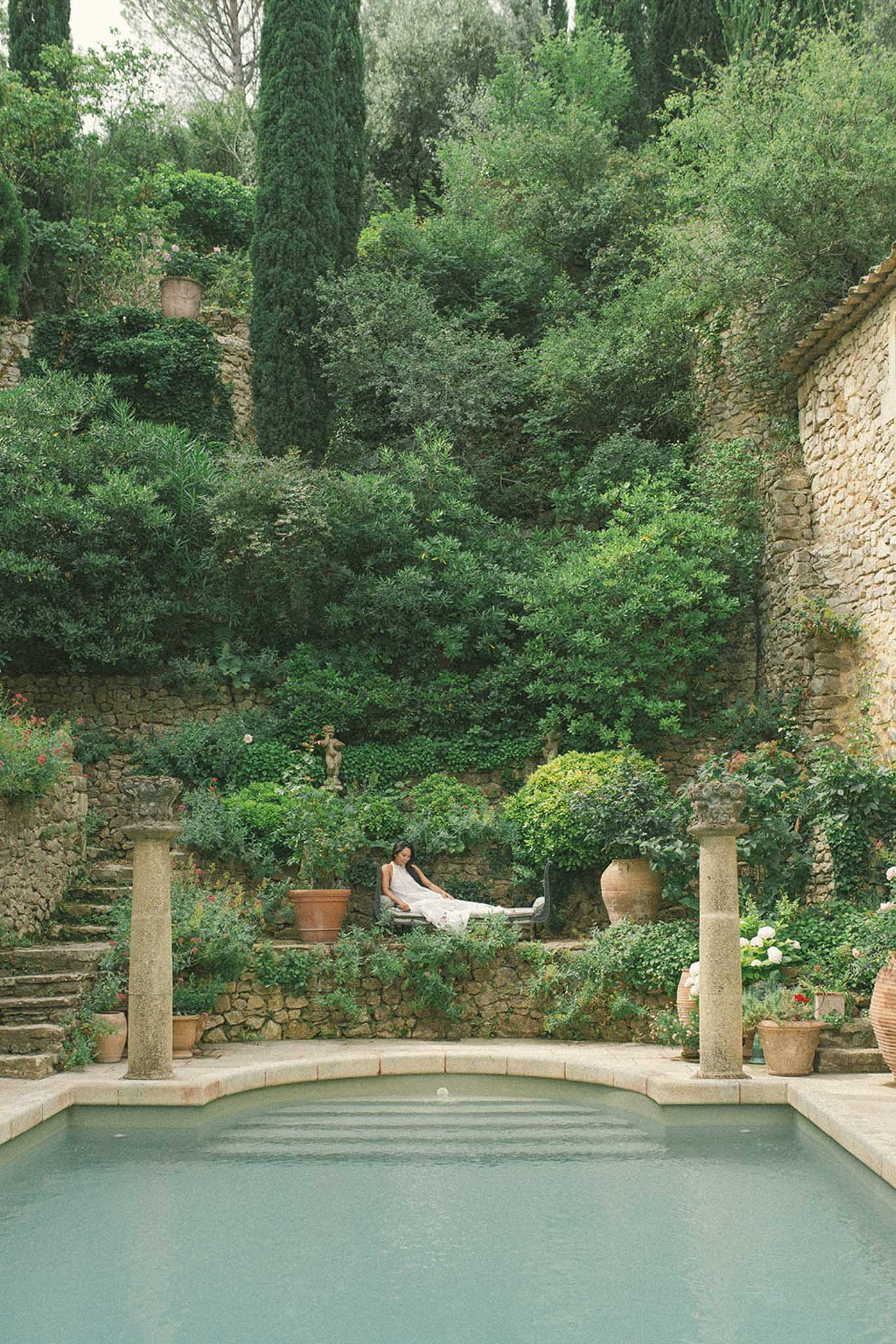 Bride reclining by garden pool surrounded by cypress trees and ivy-covered stone walls