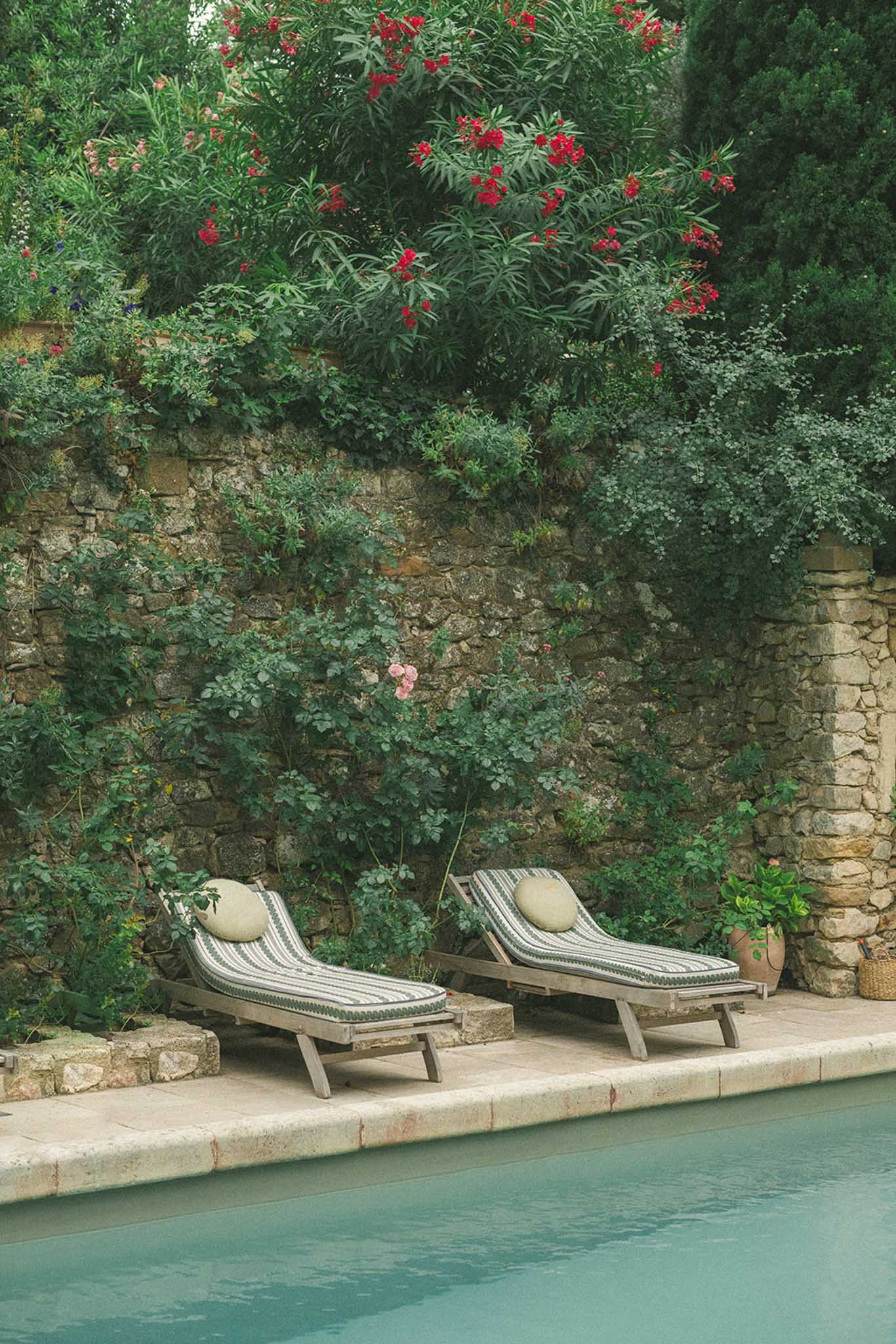 Poolside loungers with cream cushions on stone terrace beside turquoise pool and vine-covered stone walls