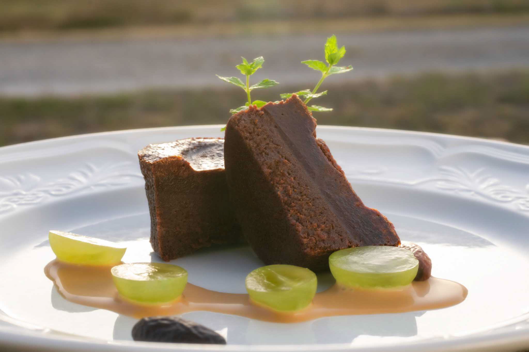 Plated chocolate dessert with mint garnish, halved grapes, and caramel sauce on white embossed plate