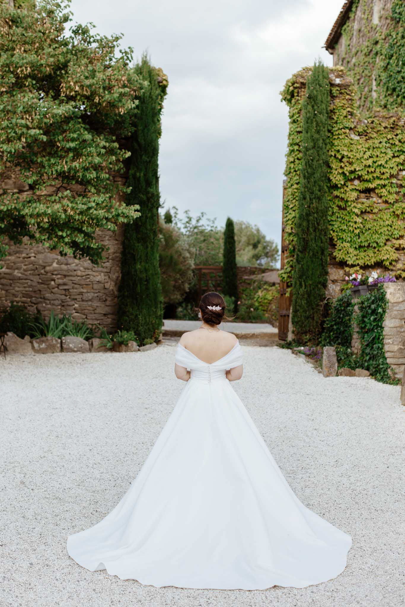 Bride from behind in off-shoulder ballgown with cathedral train spread on gravel courtyard flanked by cypress trees