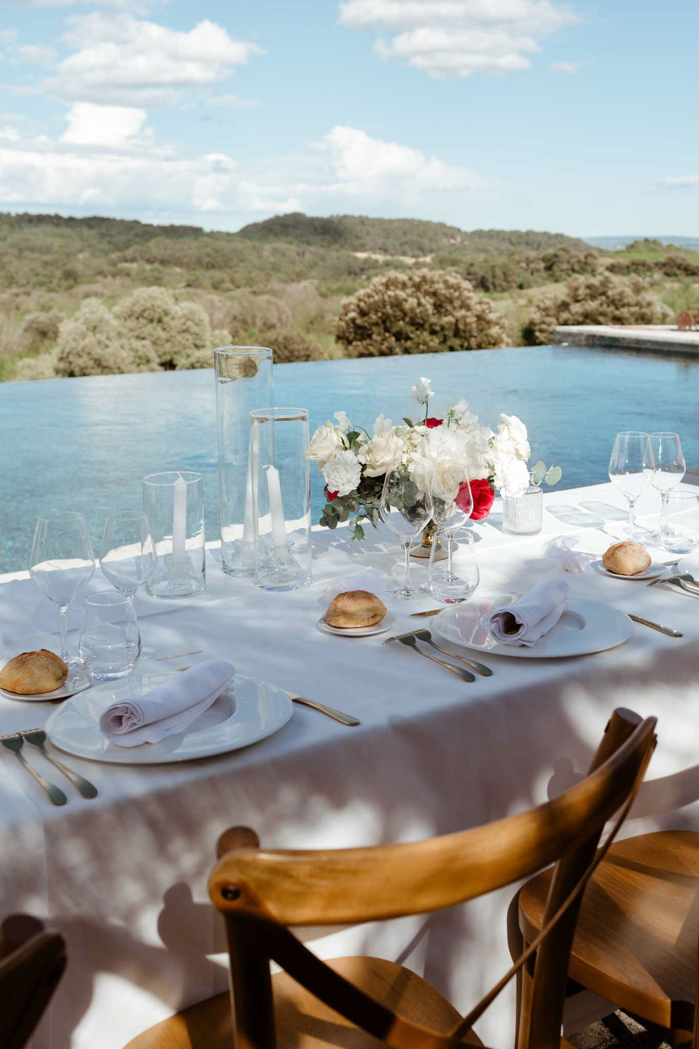 Outdoor reception table by infinity pool set with gold-trimmed plates, white and red rose centerpiece, and taper candles
