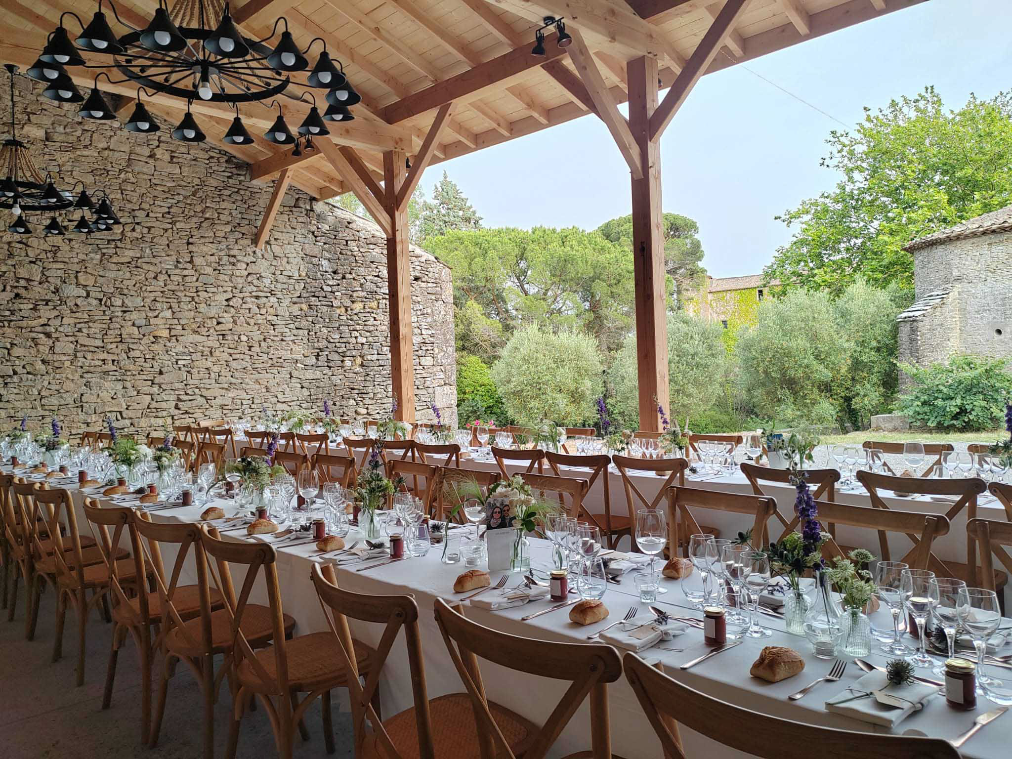 Long banquet tables set under a timber-beam pavilion with wrought-iron chandeliers and purple floral centerpieces