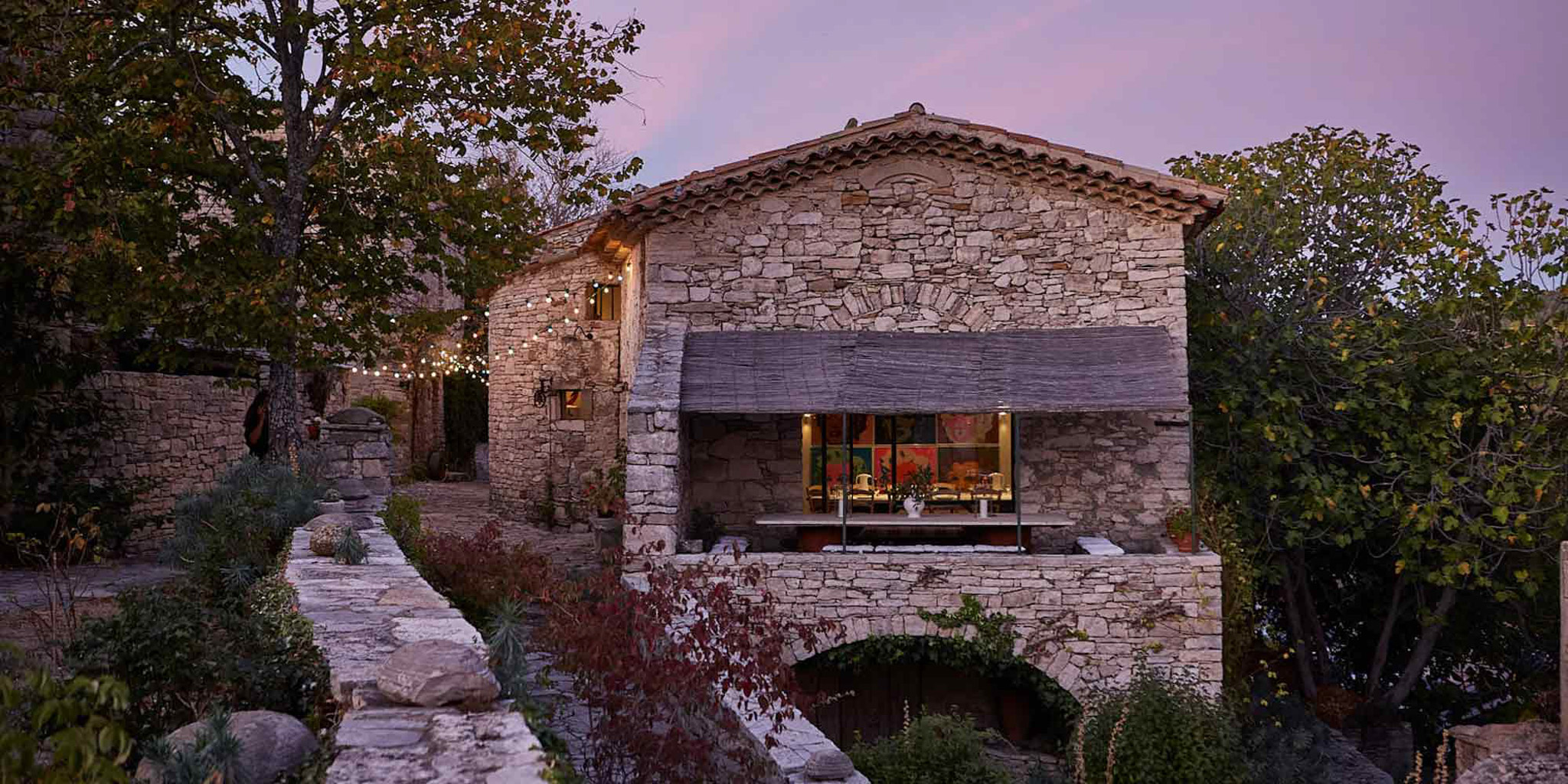 Provencal stone mas at dusk with warm interior lighting and string lights along garden pathway