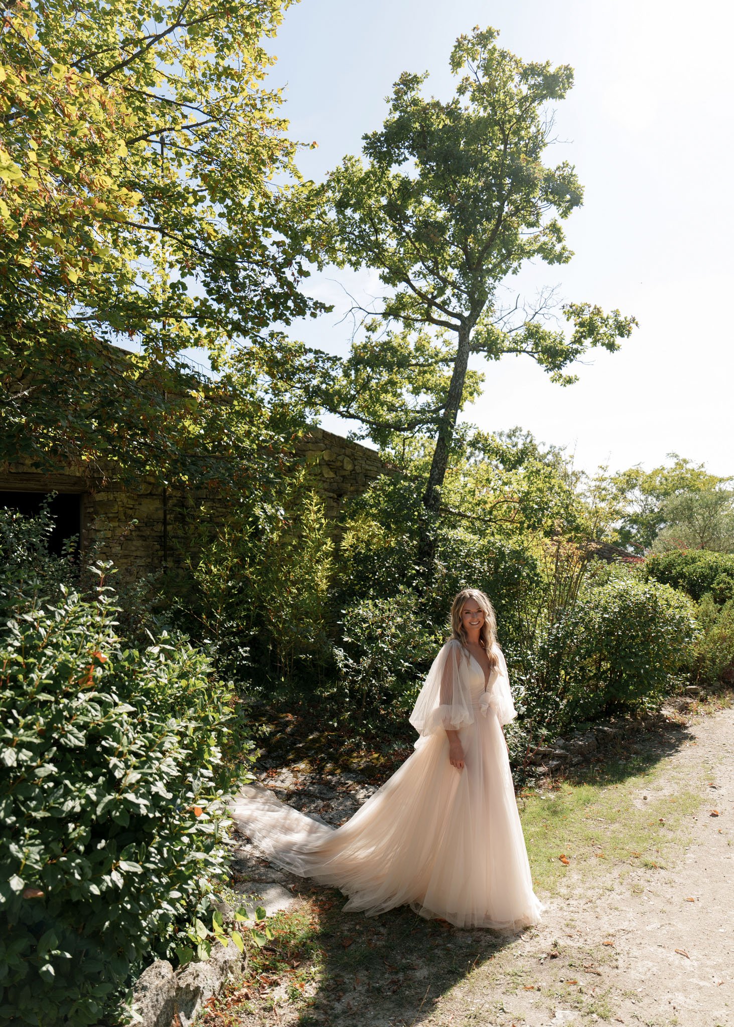 Bride in blush tulle ball gown with flutter sleeves standing on gravel path beside stone building and garden