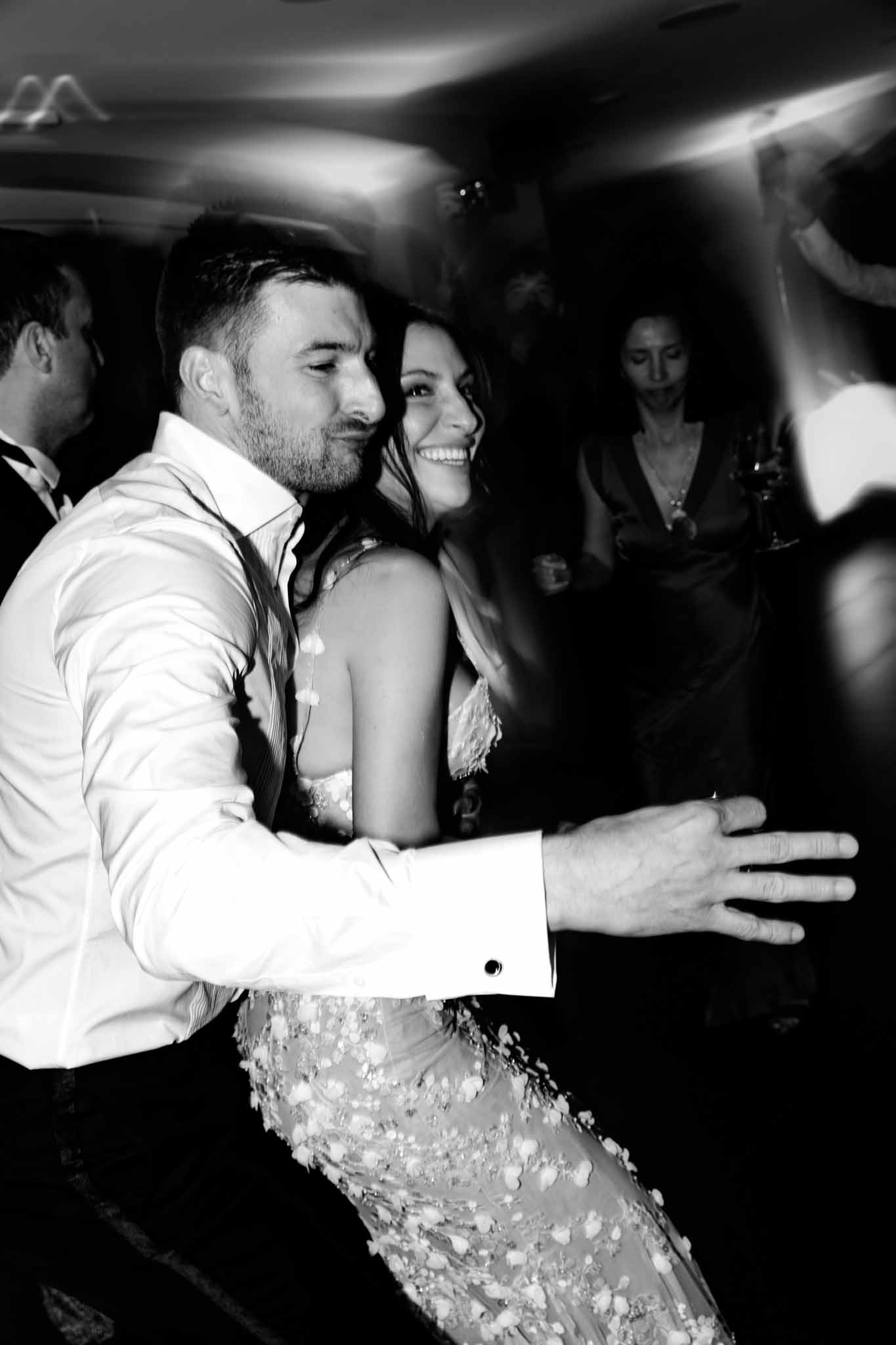 Couple dancing energetically on reception dance floor with guests behind in black and white