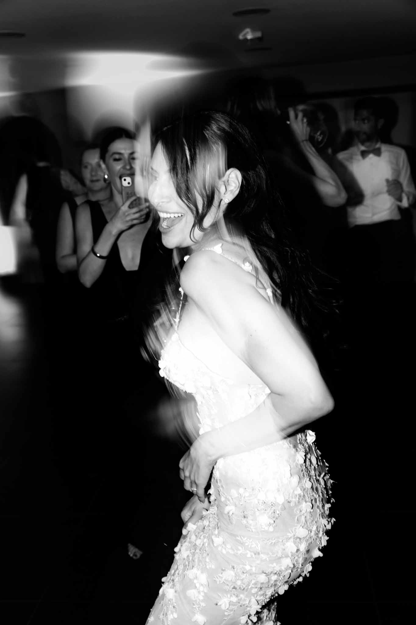This black-and-white image captures a bride dancing energetically during an indoor wedding reception. She is wearing a fitted gown with three-dimensional floral appliqué detailing and thin spaghetti straps, and her long dark hair is in motion, creating intentional blur that conveys movement and energy. Her expression shows open, joyful laughter. In the background, several guests watch and react — a woman in a dark sleeveless dress films the moment on her phone, and a man in a white dress shirt with a black bow tie looks on — both slightly out of focus. The high contrast black-and-white tones and motion blur give the image a spontaneous, candid quality; it is a mid-range portrait shot taken in what appears to be a darkened ballroom or reception hall.