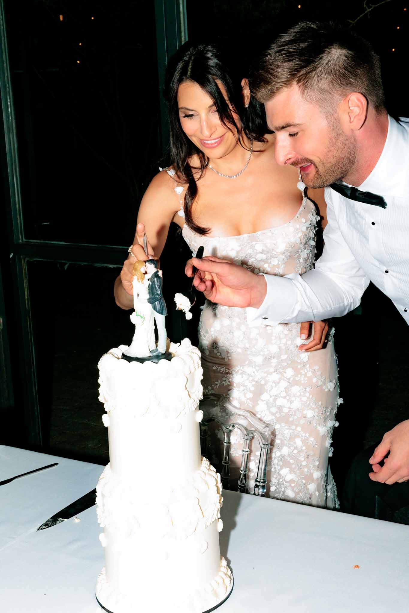Couple cutting two-tier ruffled fondant cake with figurine topper at dimly lit indoor reception