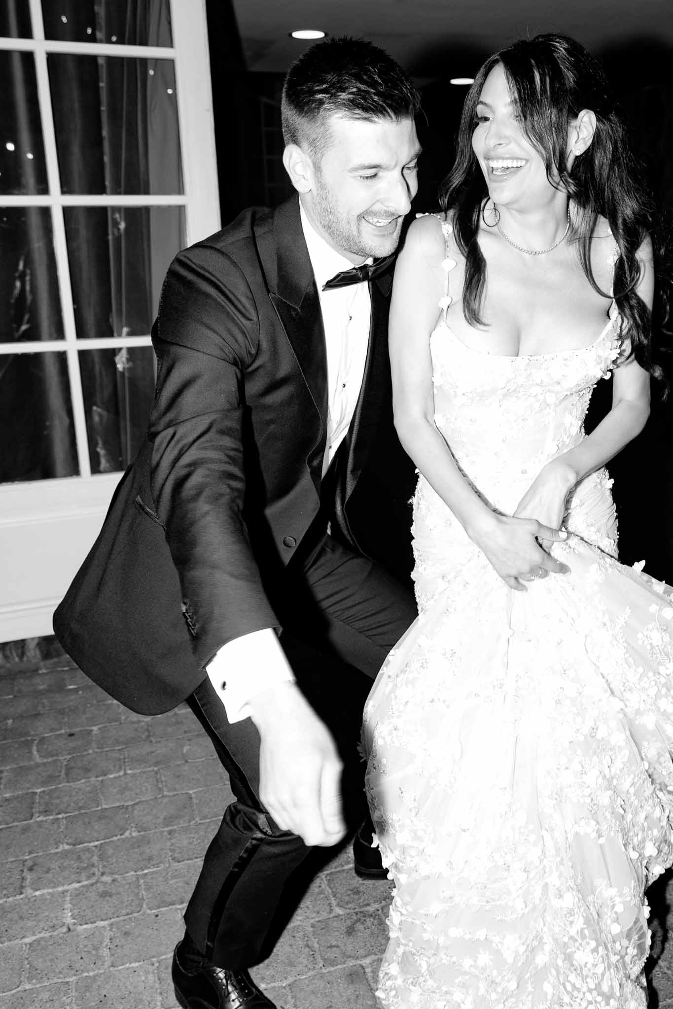 This black-and-white image captures a bride and groom dancing and laughing together during what appears to be an indoor evening reception, with a large white-framed window visible in the background. The groom wears a black tuxedo with a bow tie and satin lapels, and is caught mid-move in a playful, energetic dancing pose. The bride wears a strapless tiered ball gown with three-dimensional floral appliqué detailing throughout, accessorized with a delicate necklace and hoop earrings, with her long dark hair worn loosely down. The image has high contrast with deep blacks and bright whites, and is shot as a close-up portrait capturing the genuine, joyful expressions of both the bride and groom on a cobblestone floor.
