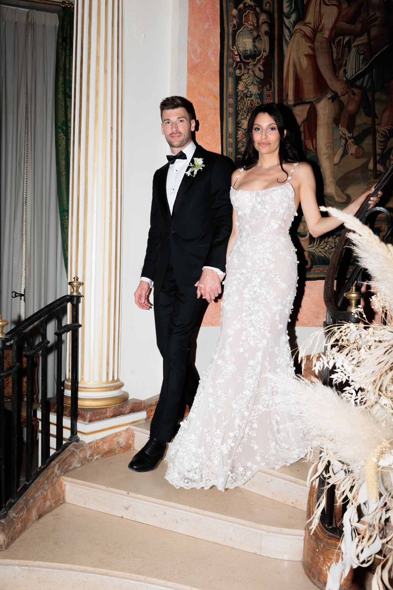 A couple poses hand-in-hand on an indoor grand staircase, likely inside a château or historic manor, captured in a mid-length portrait shot. The bride wears a fitted, semi-sheer white gown with 3D floral appliqués covering the length of the dress and thin strap detailing at the shoulders, paired with a delicate silver necklace. The groom wears a black tuxedo with a black bow tie, white dress shirt, and a small white floral boutonnière. The interior setting features gilt-accented classical columns, ornate wall-mounted tapestries with figurative scenes in terracotta and muted tones, and black wrought-iron stair railings with gold-toned newel posts. A large dried floral arrangement of pampas grass and dried foliage in warm beige and white tones is positioned at the base of the staircase to the right, contributing to a boho-classic styling mix.
