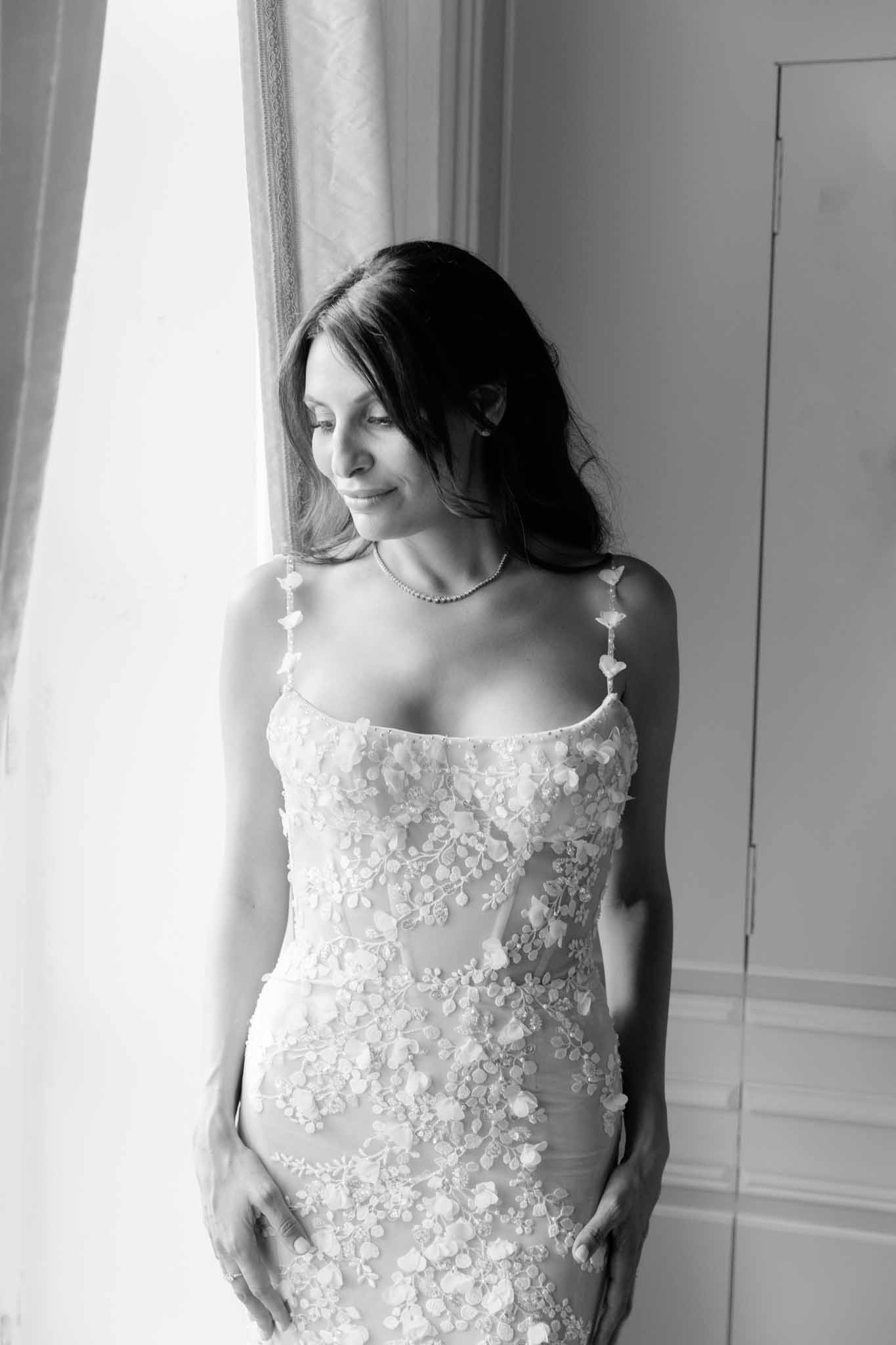 Black-and-white bridal portrait by window, bride in fitted floral applique gown with pearl necklace