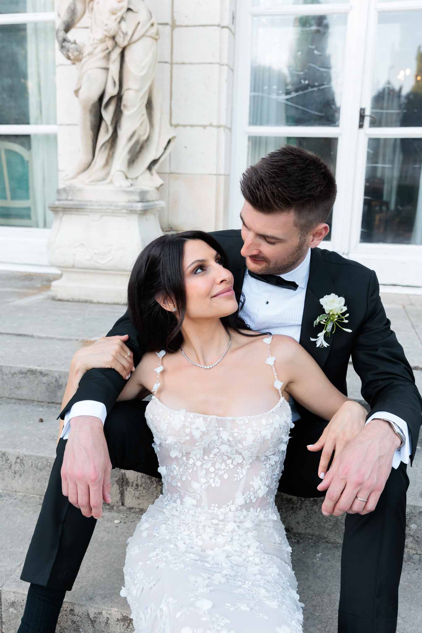 A couple portrait taken outdoors on stone steps in front of a classical French château or mansion facade, featuring large white-framed glass doors and a carved stone figurative sculpture on a pedestal. The bride, with dark shoulder-length hair, wears a fitted ivory spaghetti-strap gown with 3D floral lace appliqué detailing and a delicate diamond tennis necklace. The groom sits behind her in a black tuxedo with a black bow tie and a white rose boutonnière with green accents. He has his arms around her as she leans back against him, both looking at each other. The styling is classic and formal. Close-up portrait composition shot from a slightly elevated angle.