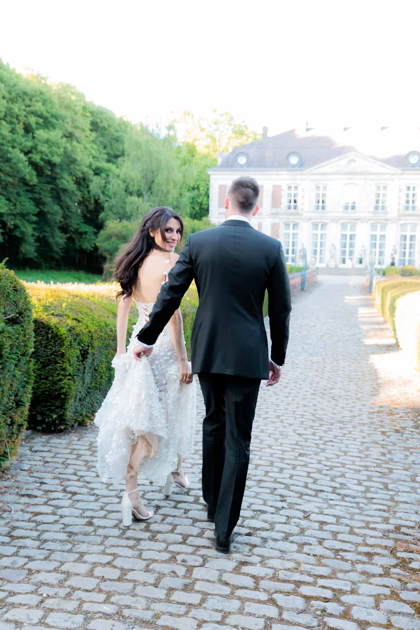 Couple walking toward French chateau on cobblestone path, bride in 3D floral applique gown with open back