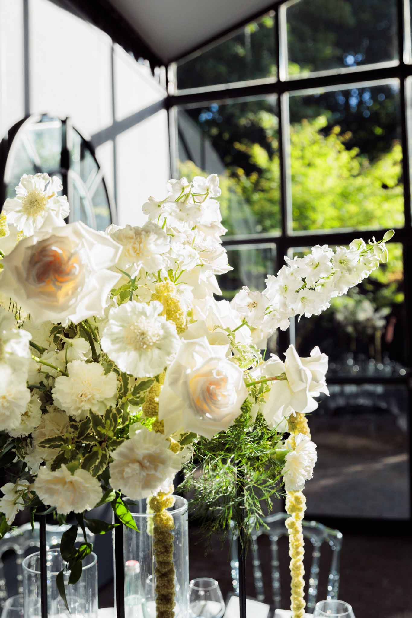 Close-up of a reception table centerpiece with white garden roses, delphiniums, anemones, and green foliage in glass vases