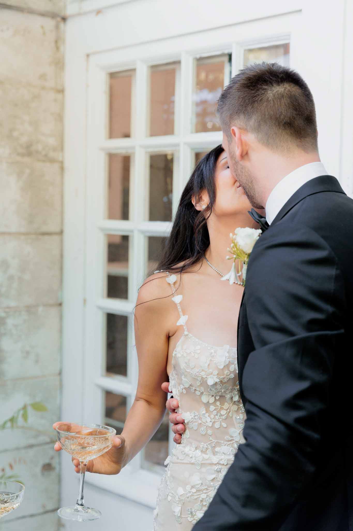 Bride in floral applique gown kissing groom holding rose champagne against chateau doors