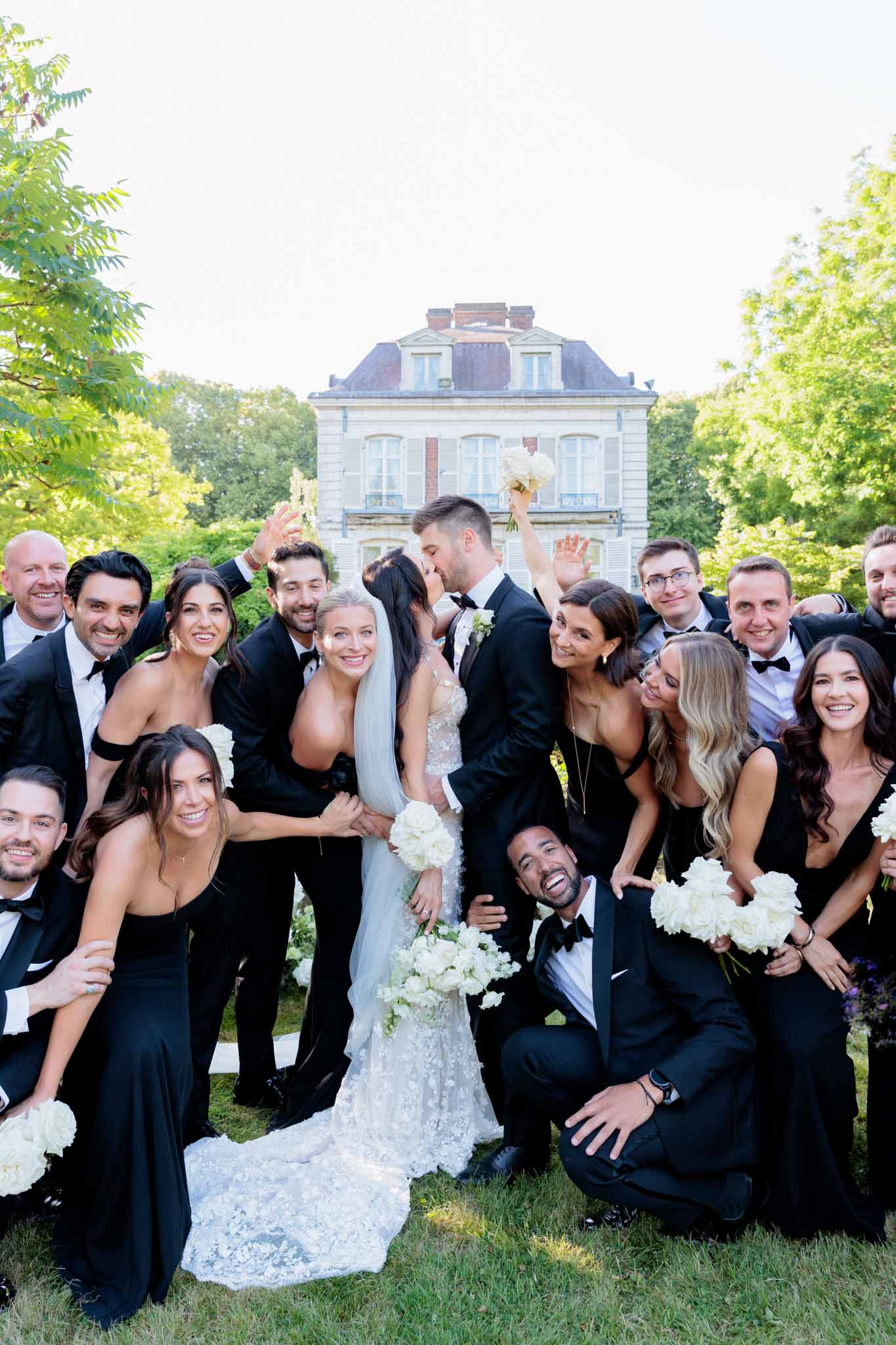 Wedding party of 16 in black gowns and tuxedos cheering as couple kisses at chateau