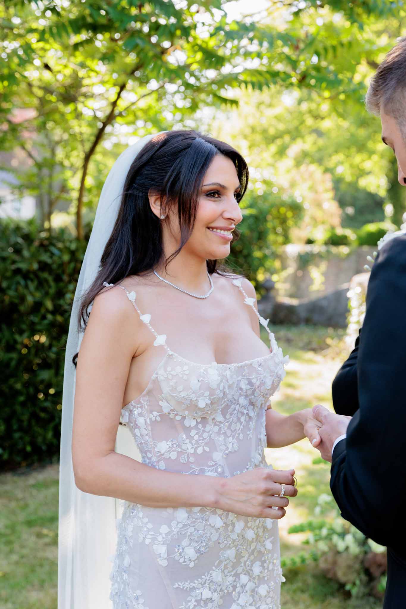 A close-up portrait of a bride during an outdoor ceremony, facing the groom whose back is partially visible on the right edge of the frame. The bride is smiling and looking toward the groom while he appears to be placing a ring on her hand. She wears a form-fitting sheer lace gown with white floral and butterfly appliqués, a plain cathedral-length veil, a delicate pearl or diamond necklace, and stud earrings. Her dark hair is worn down in loose layers. The setting is a garden with trimmed hedges and leafy trees in the background, photographed in bright natural daylight. The overall styling is classic and refined.