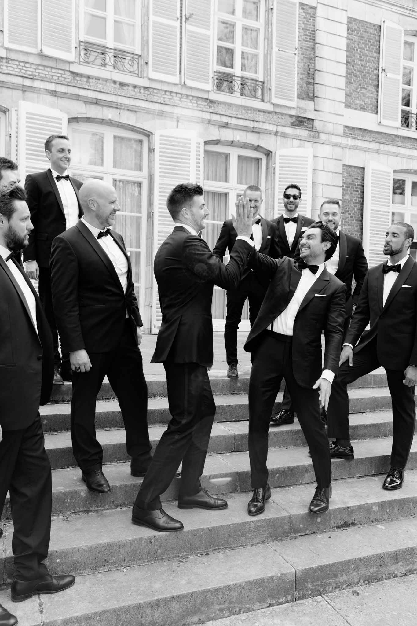 A black-and-white group portrait of the groom and approximately eight groomsmen gathered on the stone front steps of a large French manor or château with brick façade and shuttered windows. Two men in the center are giving each other a high-five, with the surrounding group reacting with laughter and smiles, creating a candid, lively moment. All men are dressed in black tuxedos with white dress shirts and black bow ties, styled in a classic formal look. The image is shot in high contrast black and white with deep shadows and bright highlights, captured as a medium-wide shot from a slight low angle.