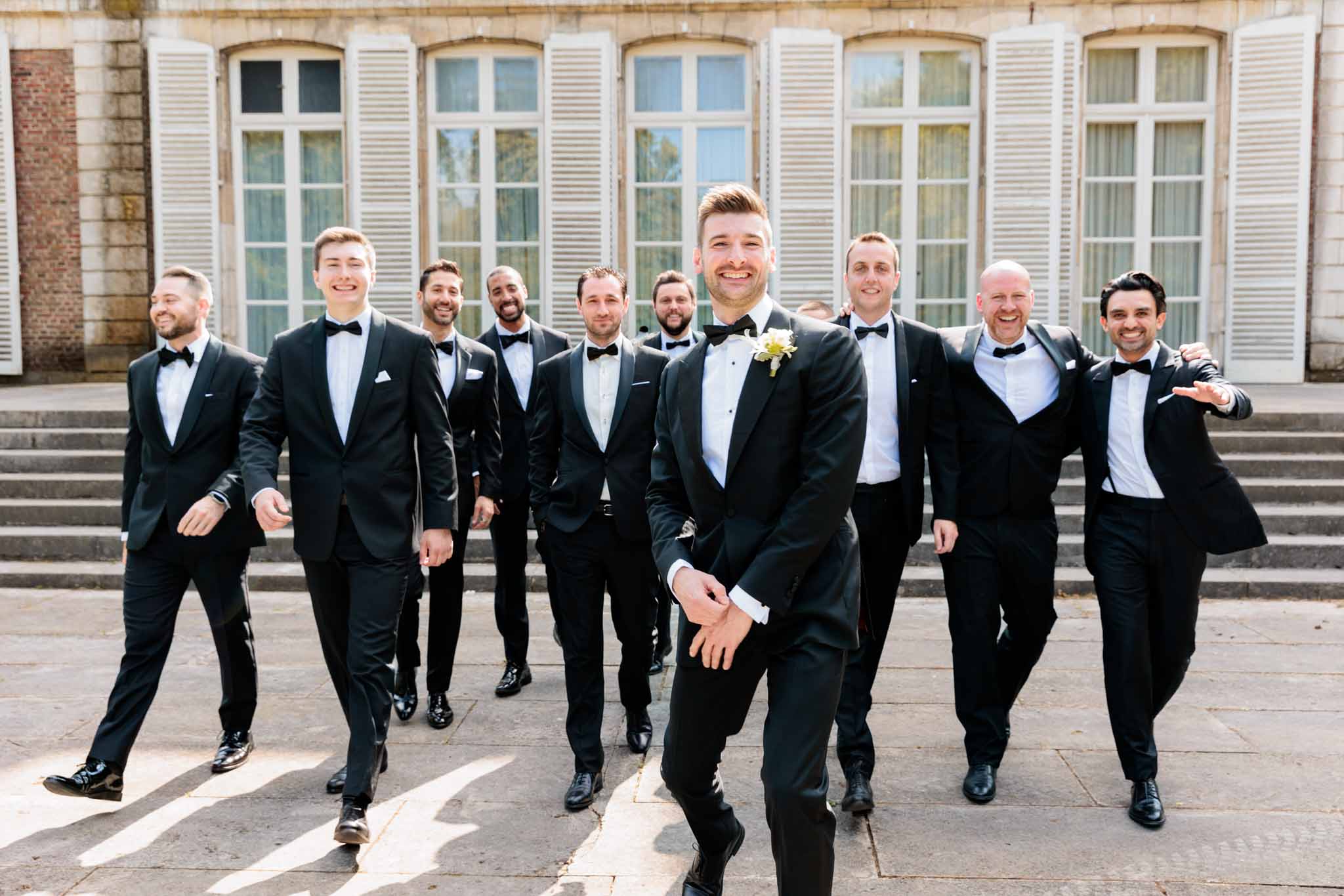 A groom and his nine groomsmen are photographed walking toward the camera on a stone-paved courtyard outside a French château or manor house, with tall arched windows and white shutters visible in the background. The groom stands in the foreground, slightly turned and smiling directly at the camera, wearing a black tuxedo with a black bow tie and a small white and yellow floral boutonnière. All groomsmen are dressed in matching black tuxedos with black bow ties and white dress shirts, some with white pocket squares; they are laughing and walking in a relaxed, candid manner. This is a wide portrait shot taken in natural daylight, with the formal classical architecture of the venue providing a structured backdrop.