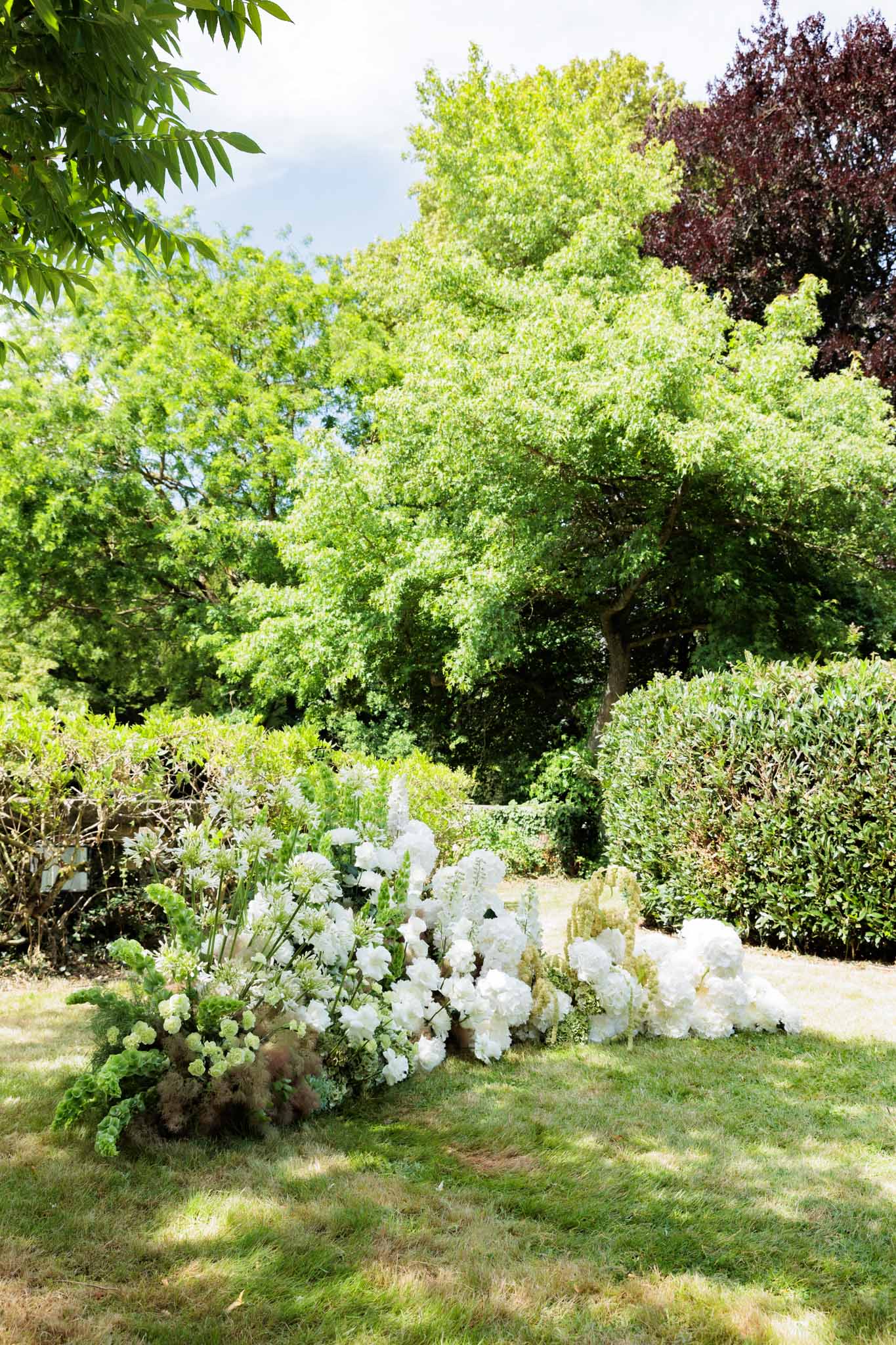 An outdoor garden setting featuring a large low floral installation arranged directly on the lawn, with no people visible. The arrangement is composed primarily of white hydrangeas, white delphiniums, white stock flowers, and green bells of Ireland, accented with soft dusty pink astilbe or similar feathery blooms and trailing greenery. The installation is horizontal and ground-level, spanning roughly two meters wide, designed in a natural, loosely structured style. The wide shot captures the floral display against a backdrop of mature garden hedging and trees, shot in full daylight.