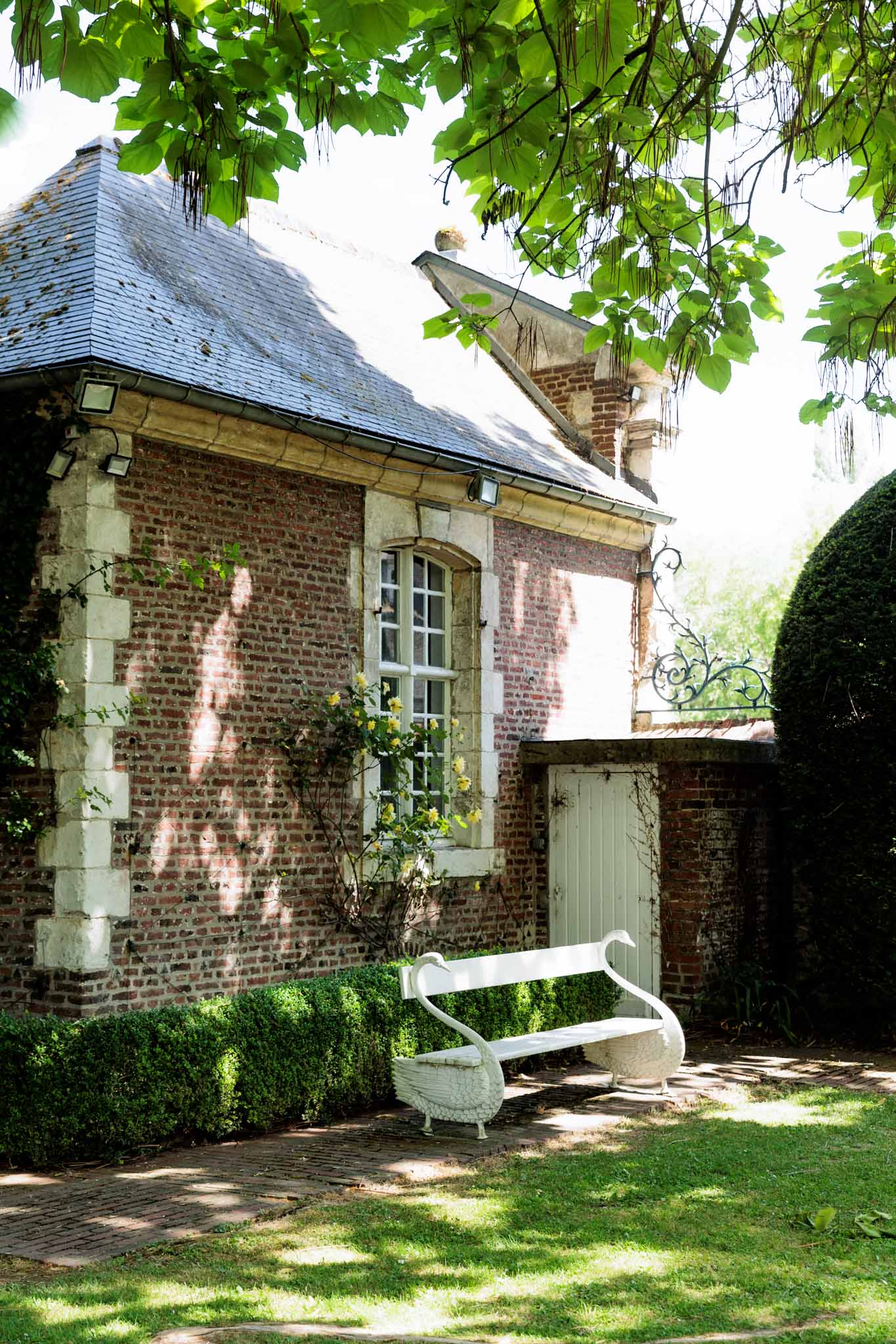An outdoor wide shot of a historic French property featuring a red brick outbuilding with white stone quoins, a slate mansard-style roof, and multi-pane arched windows. A decorative white iron bench with swan-shaped armrests sits in front of the building, flanked by a neatly trimmed low boxwood hedge. A pale sage-painted wooden door leads to a walled passage, and an ornate wrought iron gate with scrollwork is visible to the right alongside a clipped topiary sphere. No people are present in the image. Potential venue feature image.