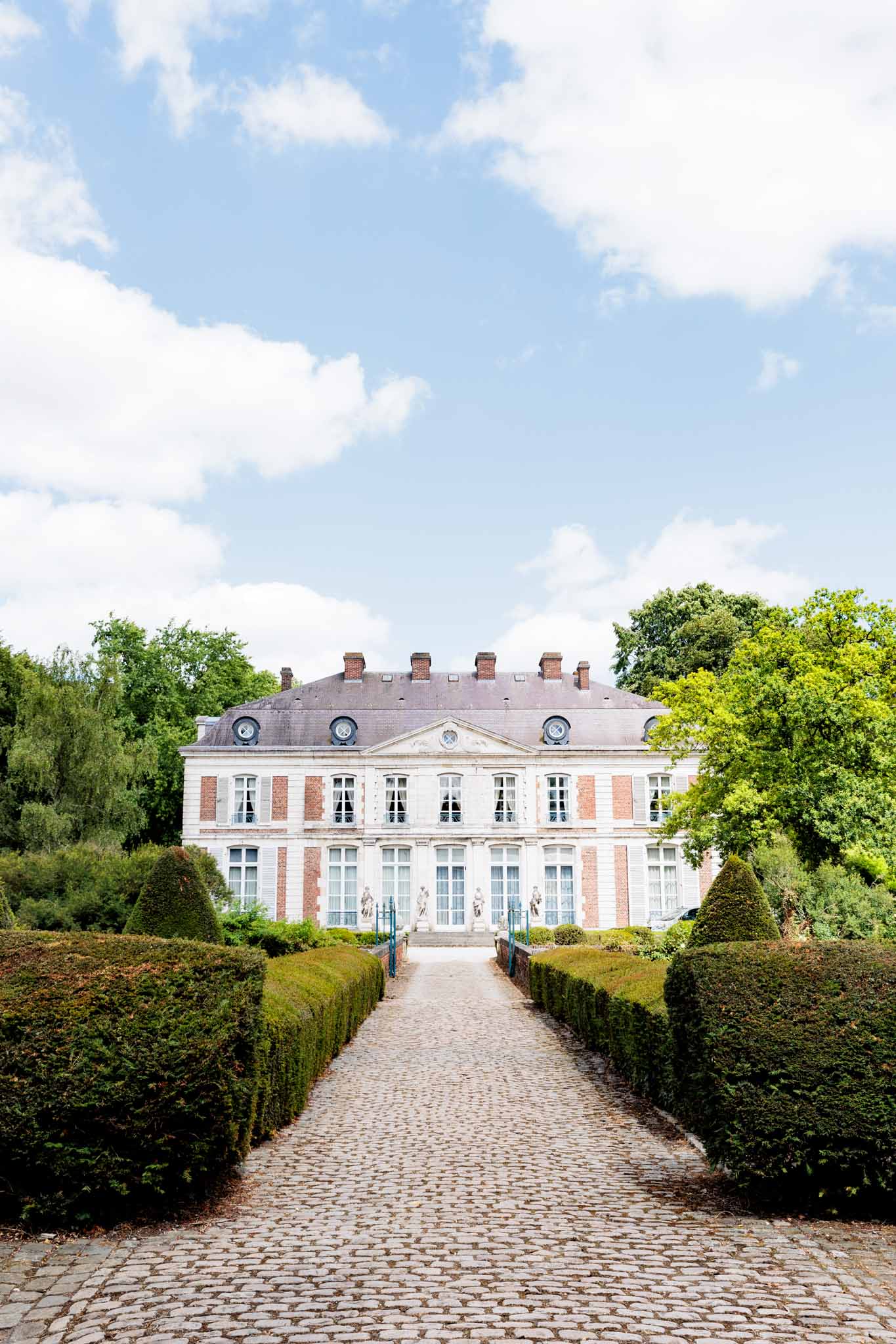 An exterior wide shot of a French château featuring a symmetrical brick and stone facade with white-trimmed tall windows across three floors, a mansard-style slate roof with multiple chimneys, and classical stone statuary flanking the entrance steps. The foreground shows a straight cobblestone pathway leading directly to the entrance, bordered by neatly trimmed low boxwood hedges and cone-shaped topiaries on either side, forming a formal French garden approach. No people are visible in the image. Potential venue feature image.
