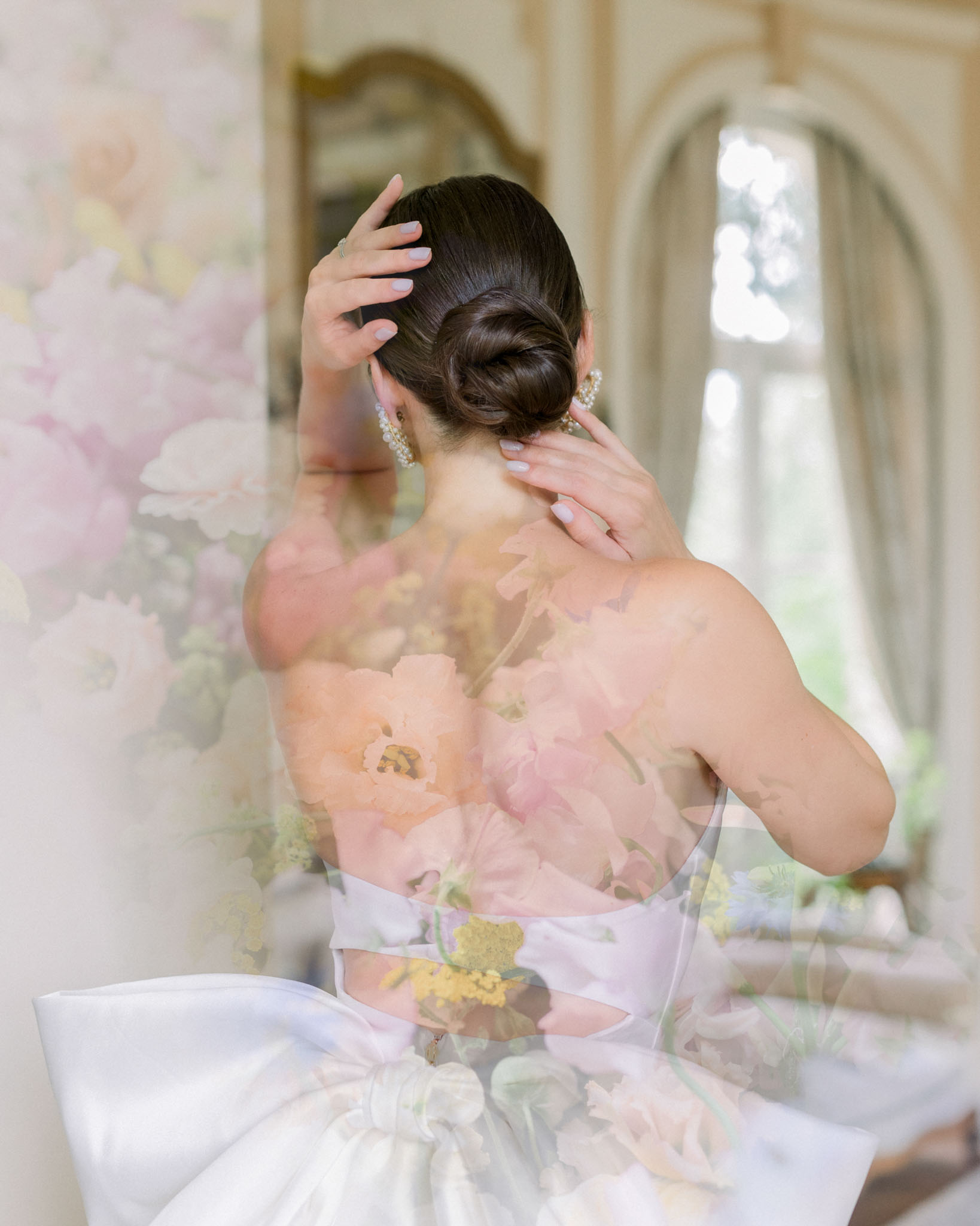 Bride from behind in strapless gown with lavender ribbon sash and pearl pins, blurred pink and white floral backdrop
