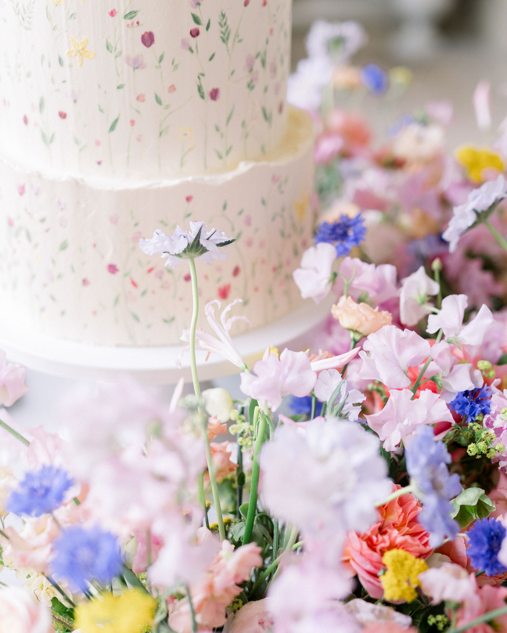 Wedding cake with hand-painted watercolor florals surrounded by pink sweet peas, blue cornflowers, and coral dahlias