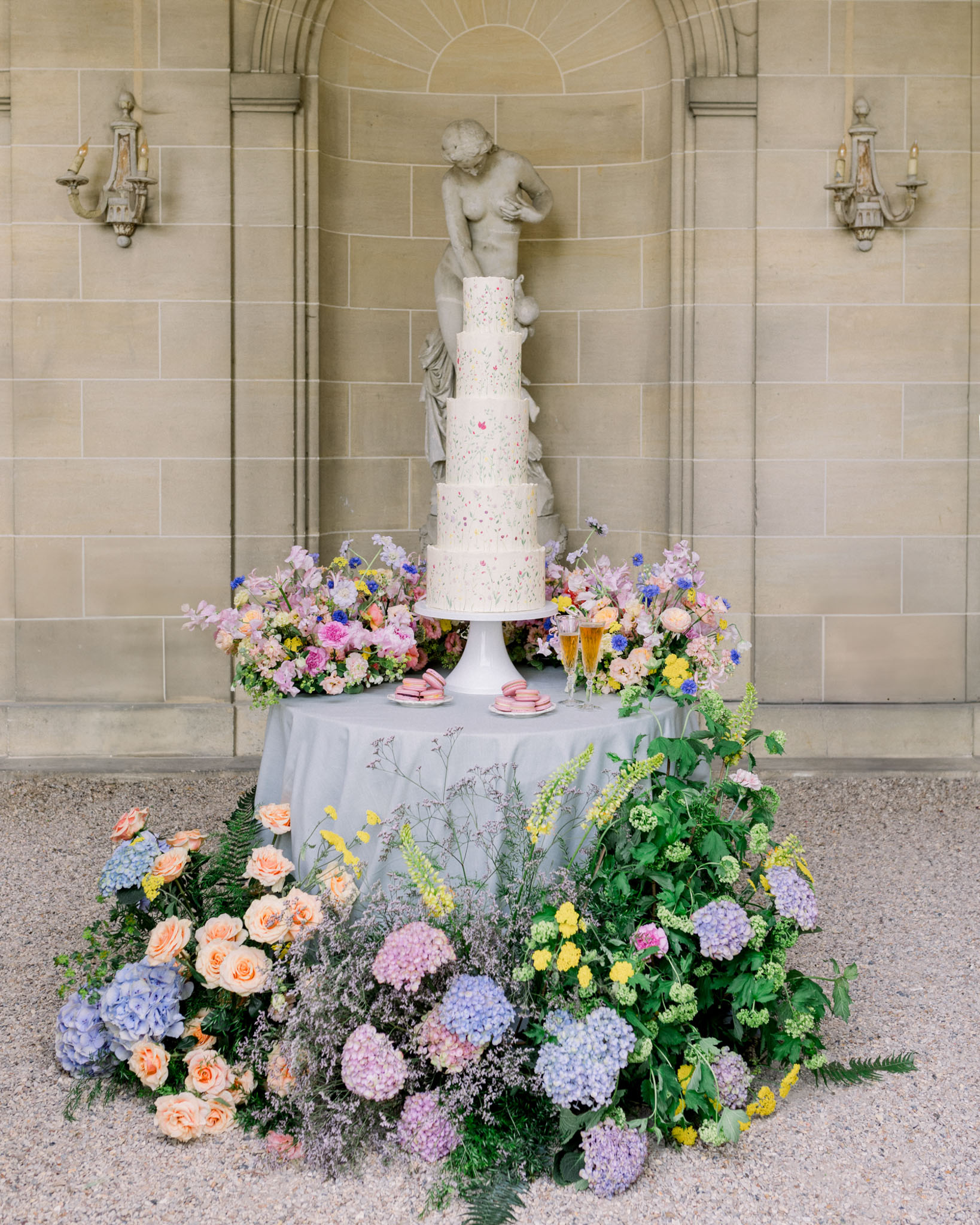 Cream wedding cake with cherub topper surrounded by cascading pastel garden flowers in stone-vaulted neoclassical venue
