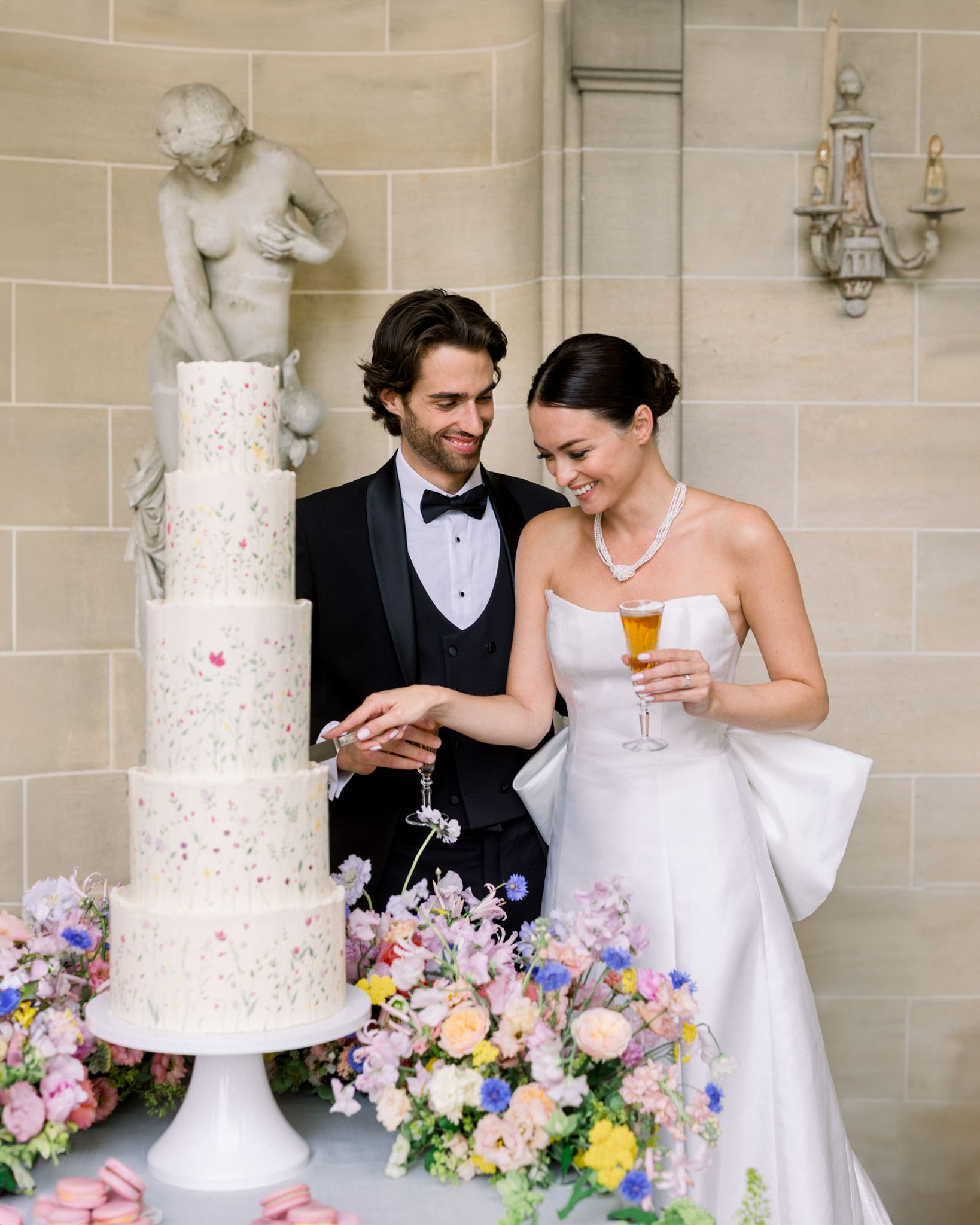 Bride and groom cutting a three-tiered ivory cake decorated with pastel florals in pink, yellow and blue tones; marble bust visible behind.