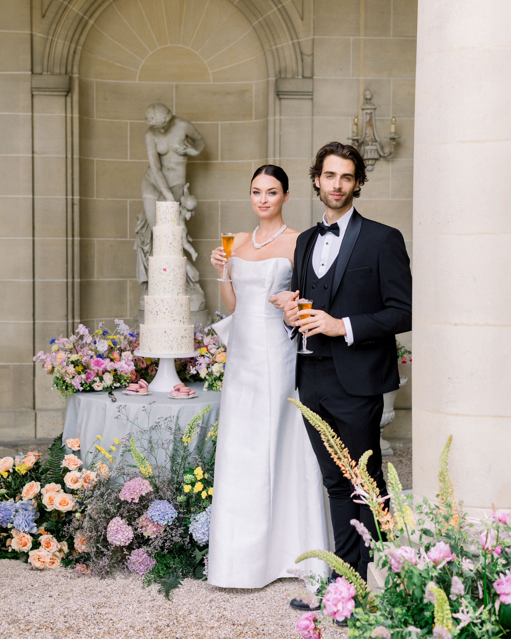 Bride and groom by wedding cake with peach roses and blue hydrangeas in neoclassical courtyard