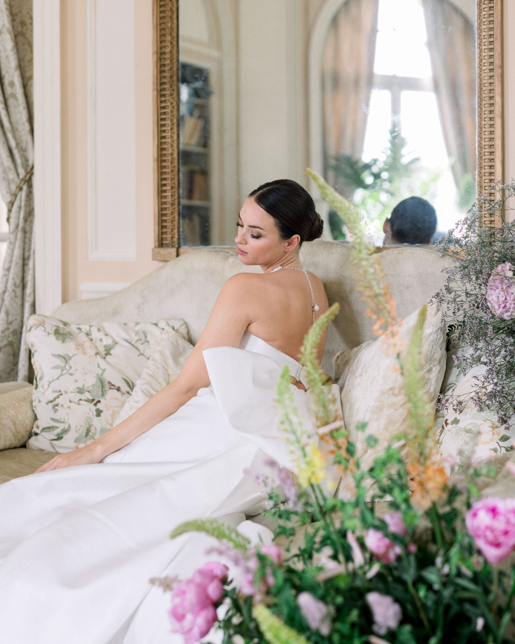 Bride seated on sofa in classical interior with pink and white flowers, gold-framed mirrors, and green drapes
