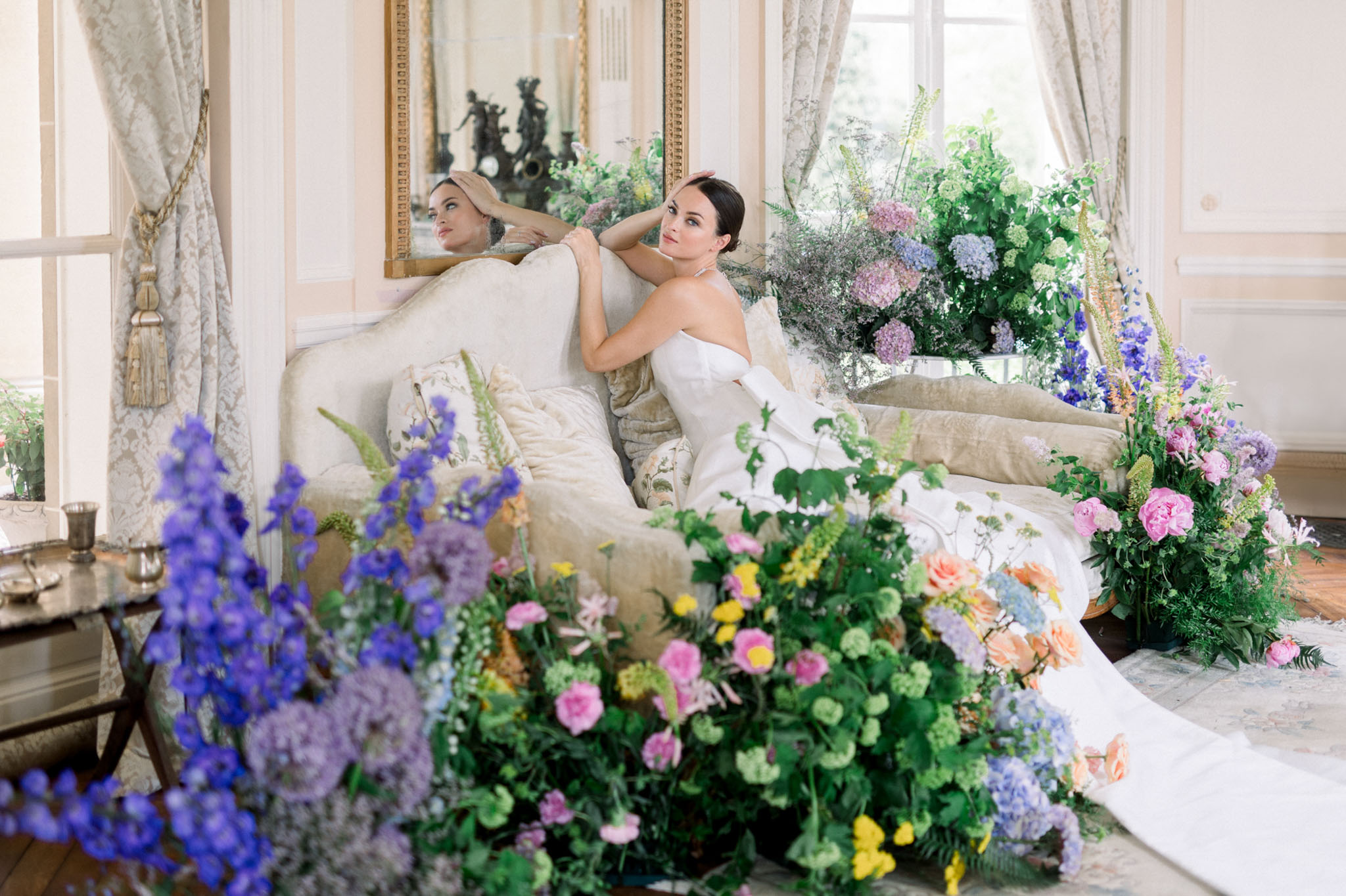 Bride in strapless white dress reclining on cream bed surrounded by purple delphiniums, pink roses, and green hydrangeas in ornate room