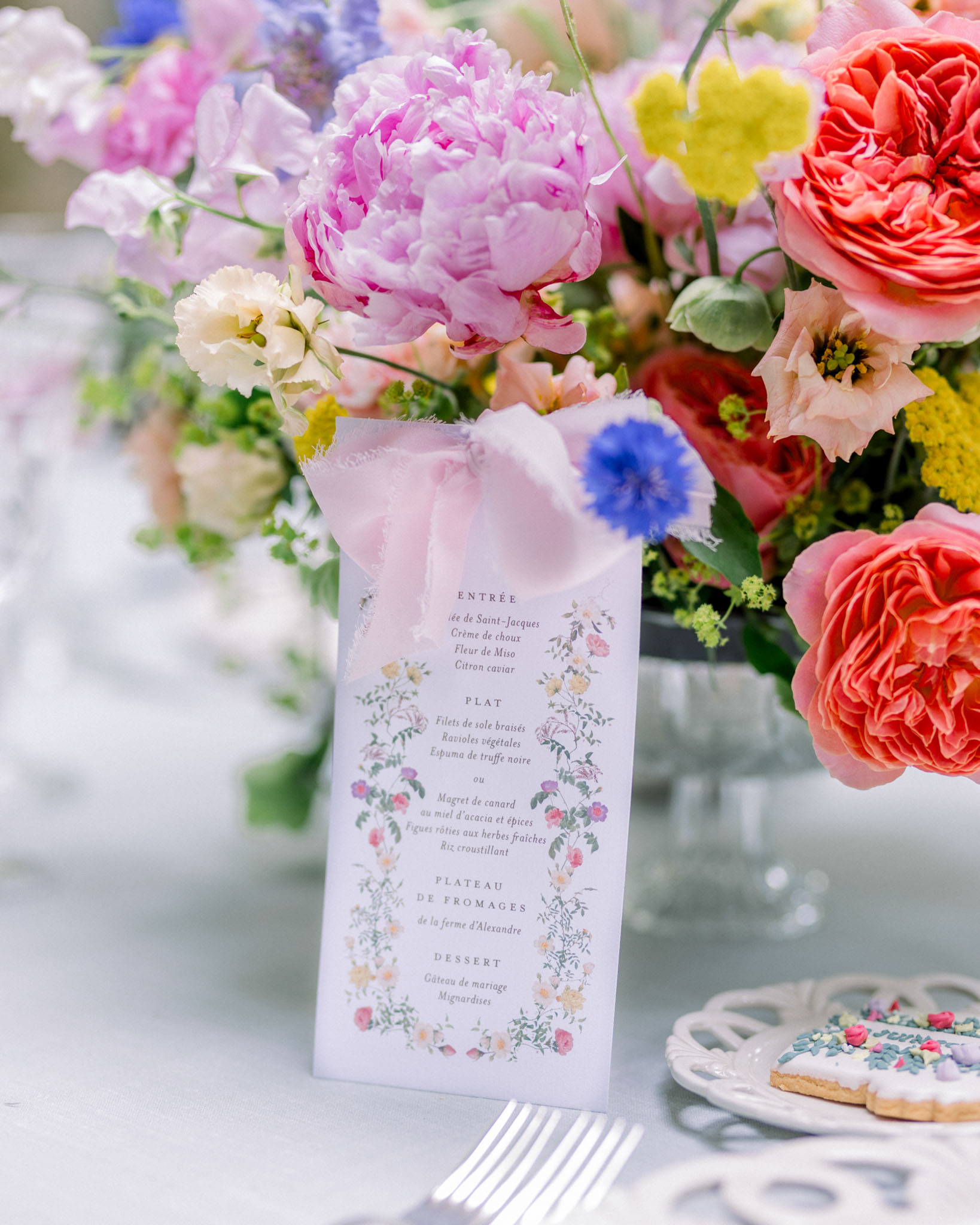 French wedding menu card surrounded by pink peonies, coral roses, blue cornflowers, and decorated cookies