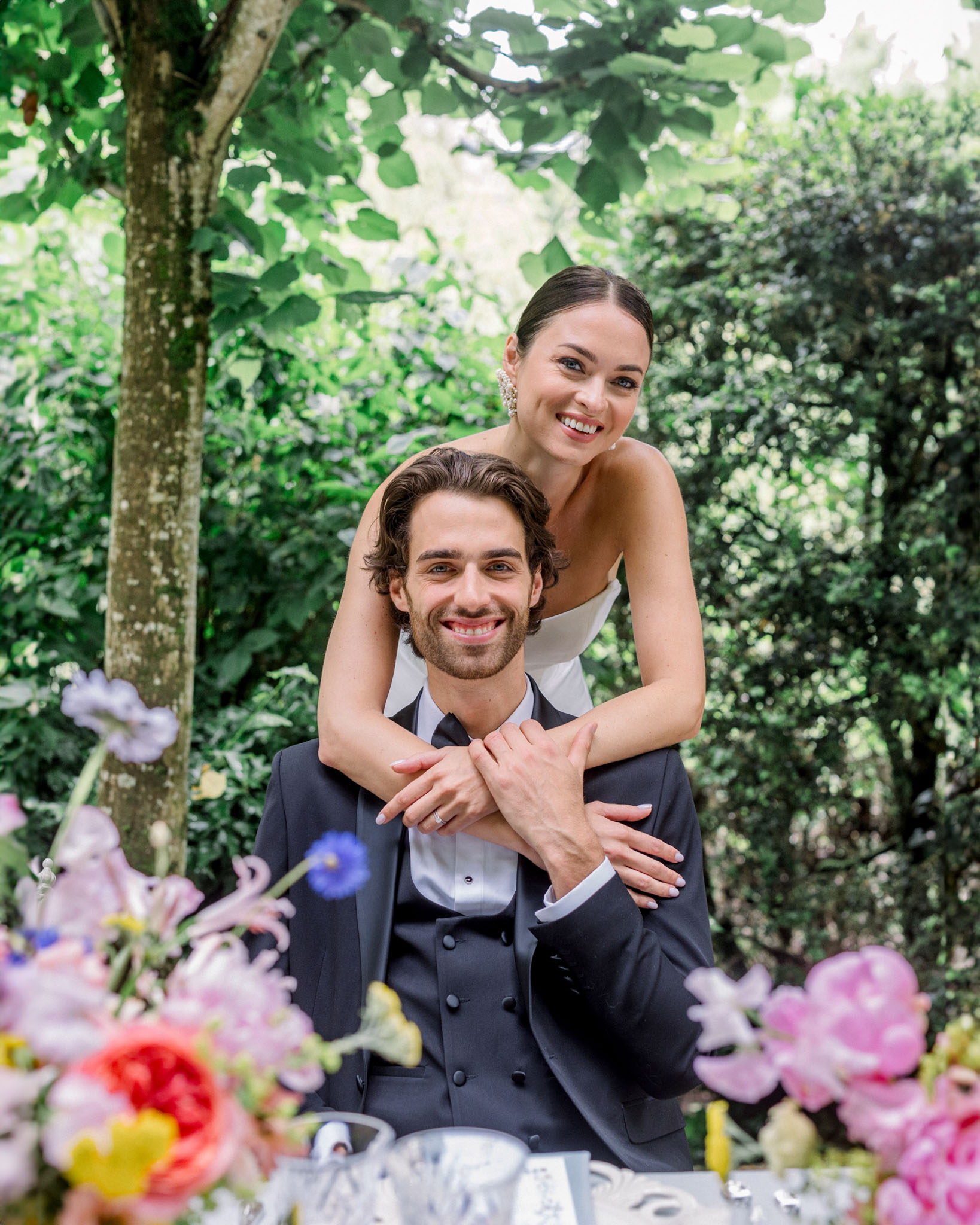 Groom in navy double-breasted suit and bride in ivory dress pose in garden with red roses and delphiniums foreground