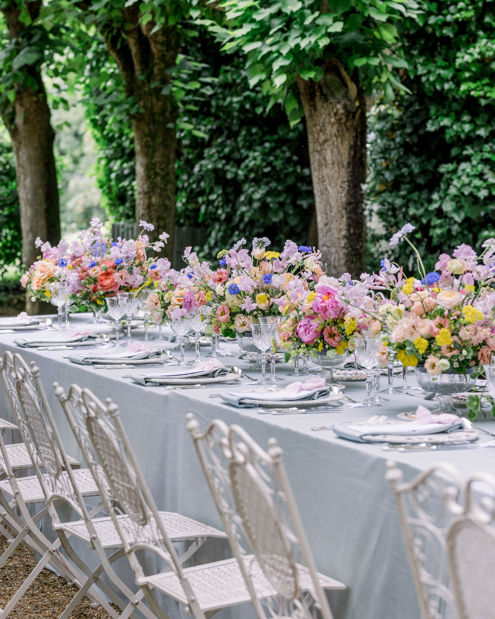 Long outdoor reception table with pale grey linen, crystal vases of garden roses and mixed florals under mature trees
