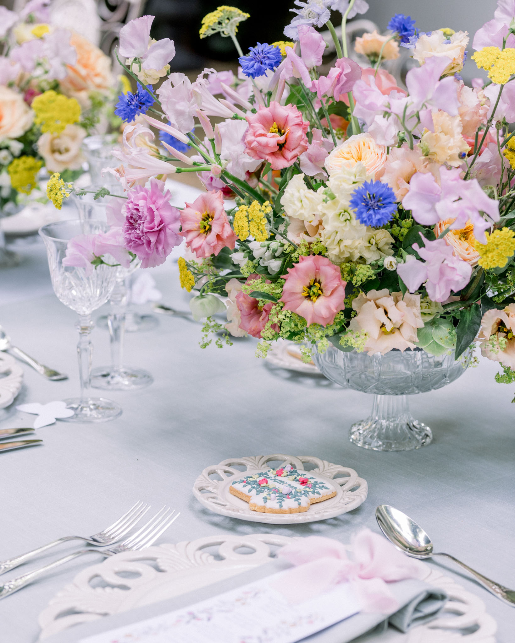 Wedding tablescape with pink peonies, coral roses and blue cornflowers in pedestal bowl with white linen