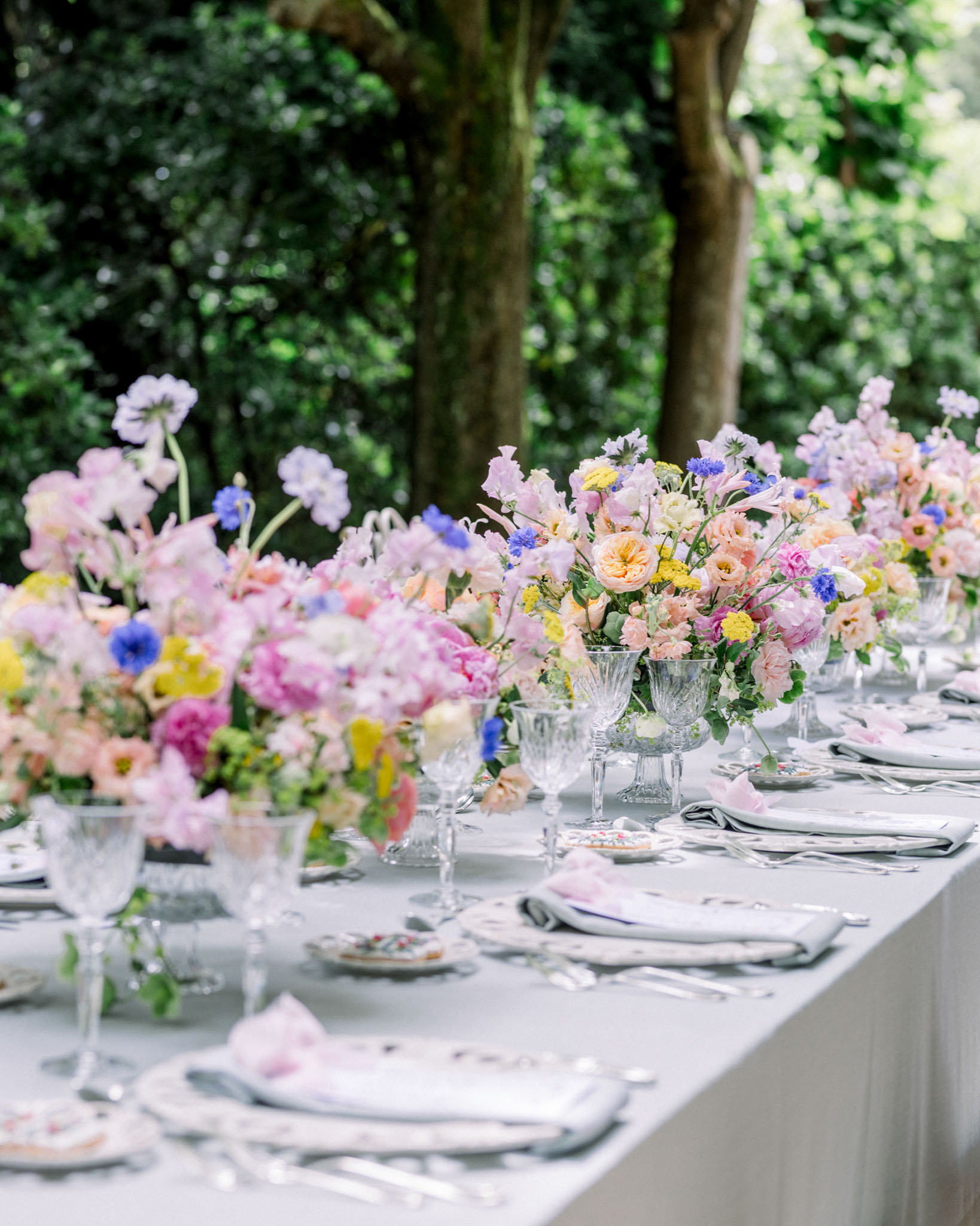 Long outdoor dining table with crystal urn vases of garden roses, cornflowers, and blue blooms under green tree canopy
