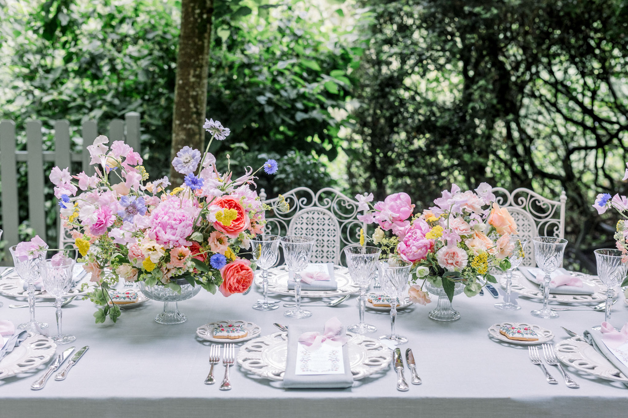 Outdoor reception table with pink peonies, coral roses, and blue delphiniums in glass compote vases, white linens and crystal glassware