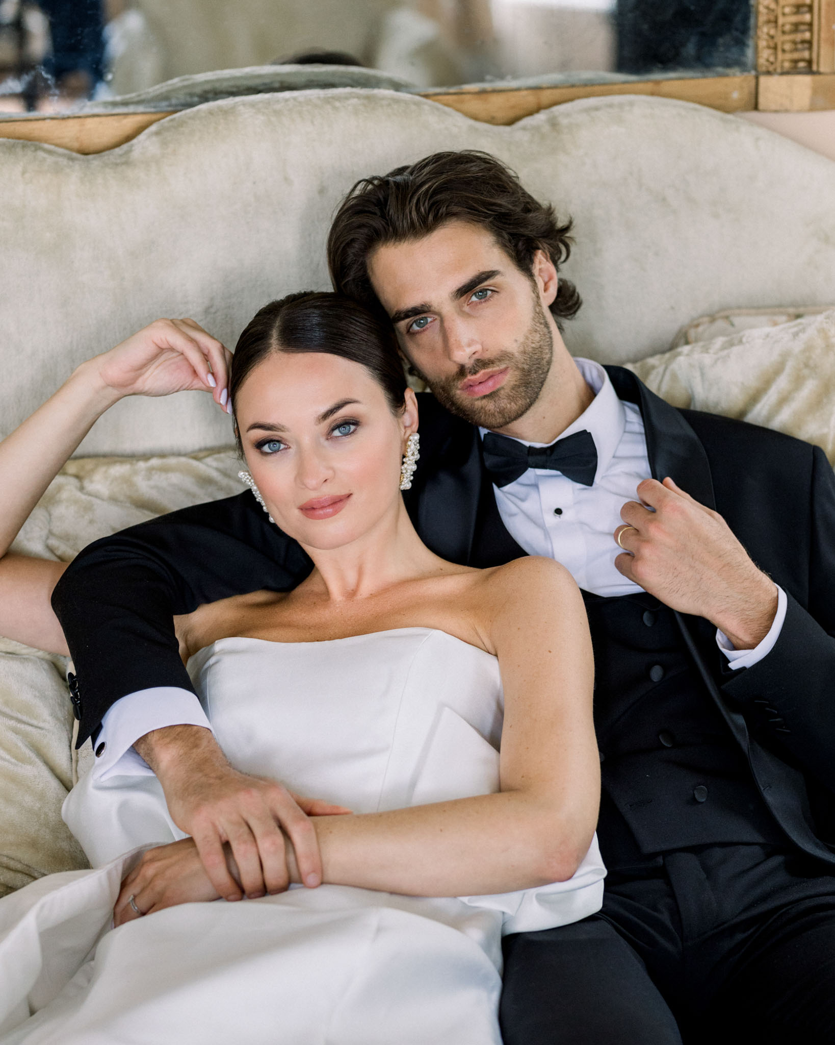Bride in strapless white dress and groom in black tuxedo seated together on cream upholstered sofa