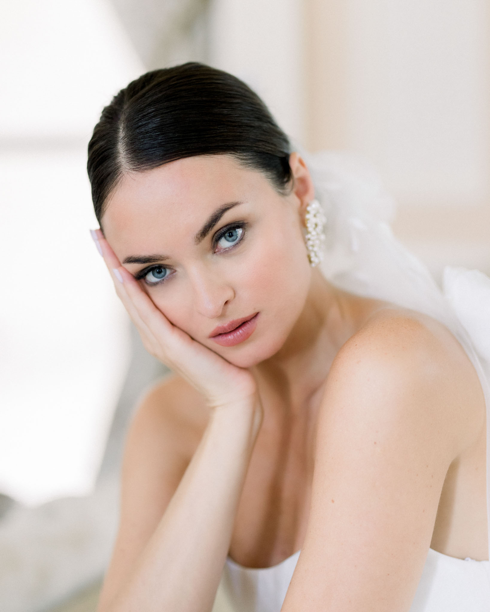 Close-up portrait of bride in strapless white dress and pearl earrings resting her chin on her hand