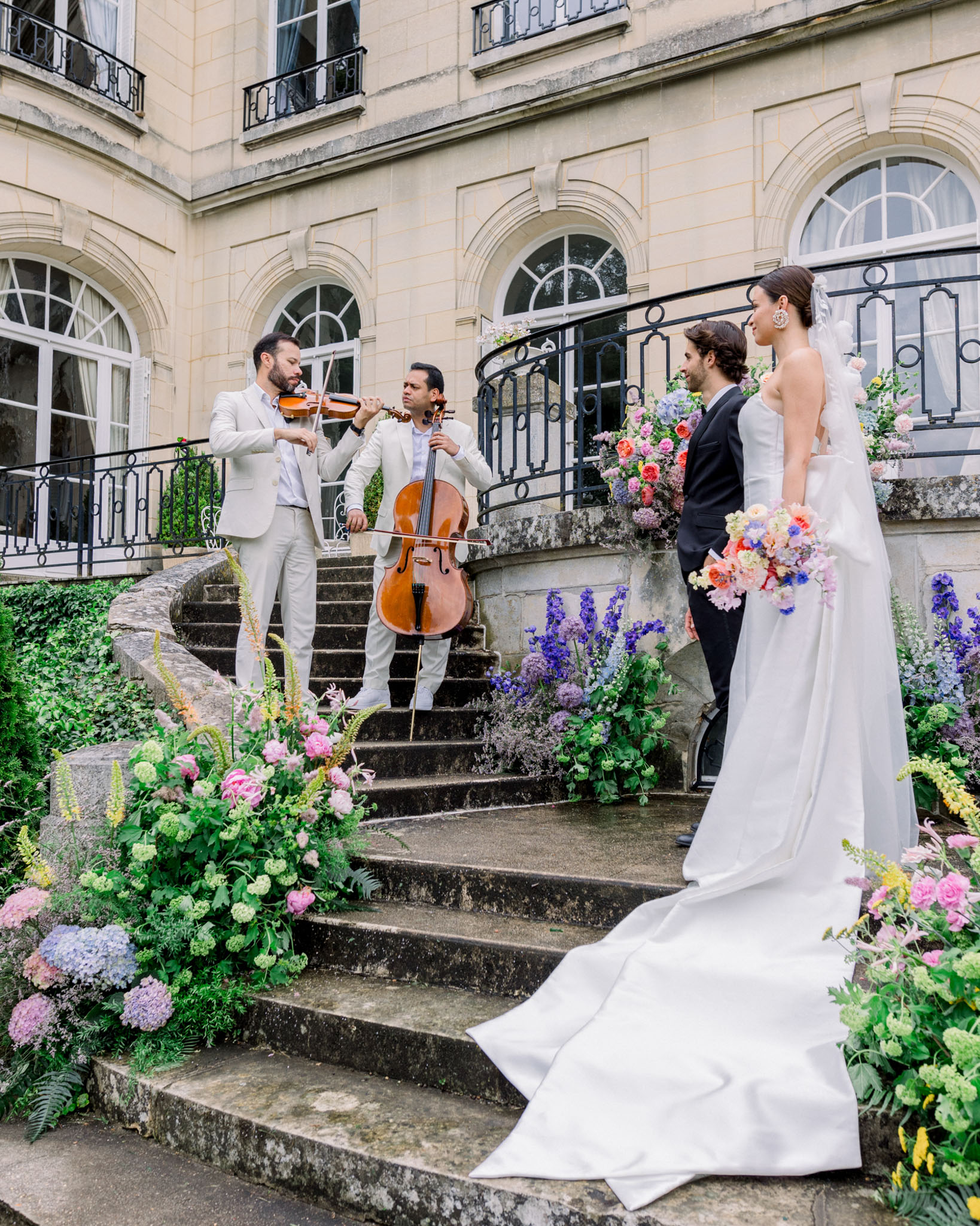 String duo on grand stone staircase, bride in ivory column gown nearby, flanked by blush and coral floral arrangements