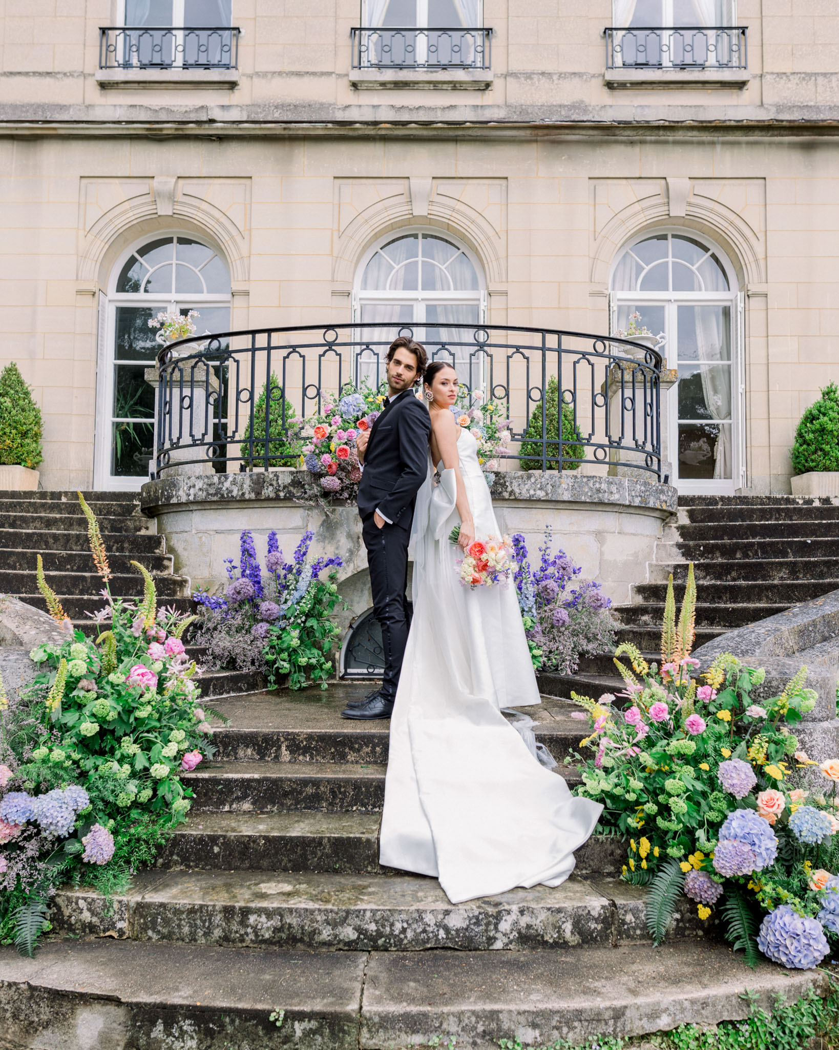 Bride with coral-pink bouquet and groom on stone château stairs flanked by abundant hydrangea and rose floral arrangements