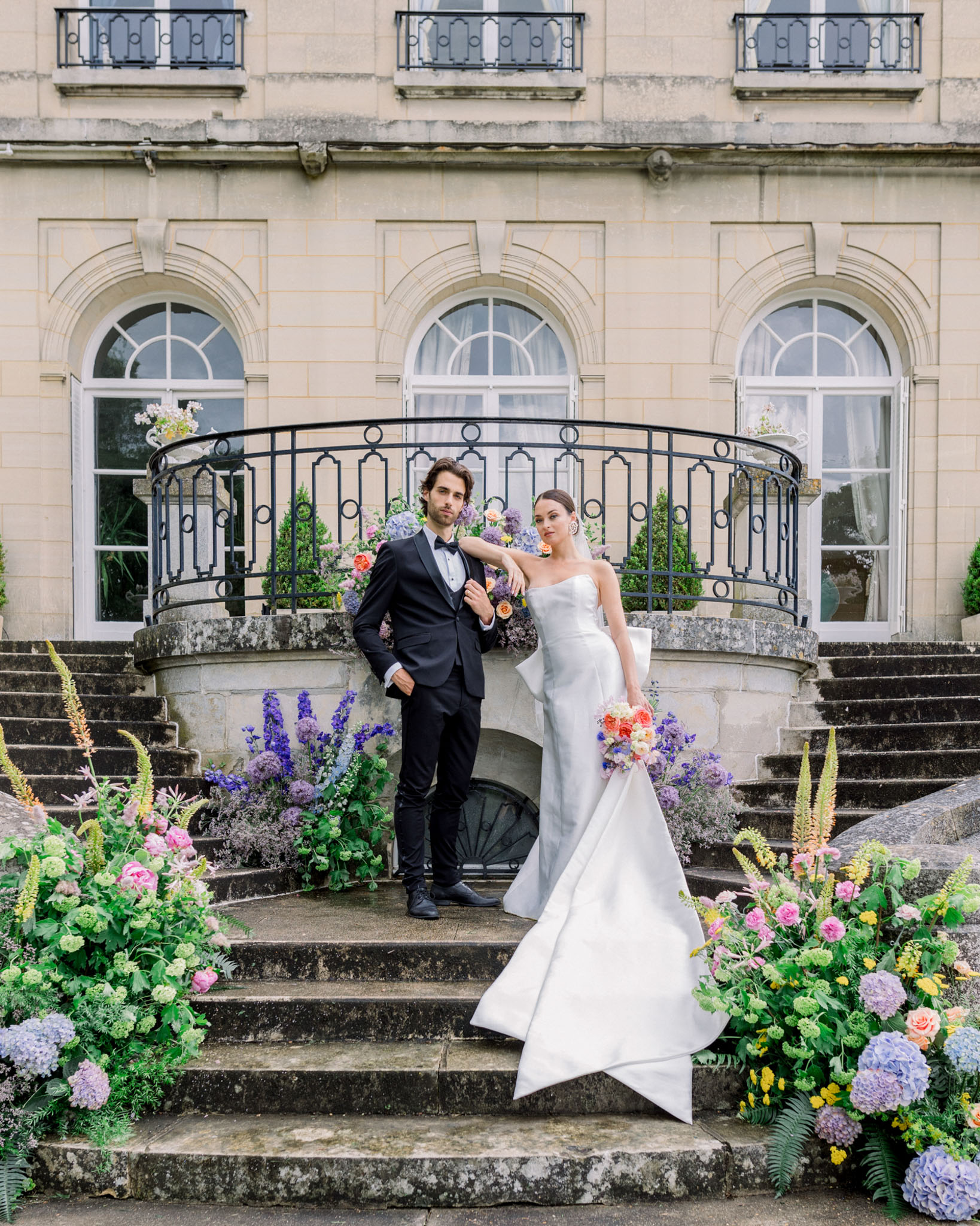 Bride and groom posing on grand stone staircase lined with coral roses and purple hydrangeas at French chateau