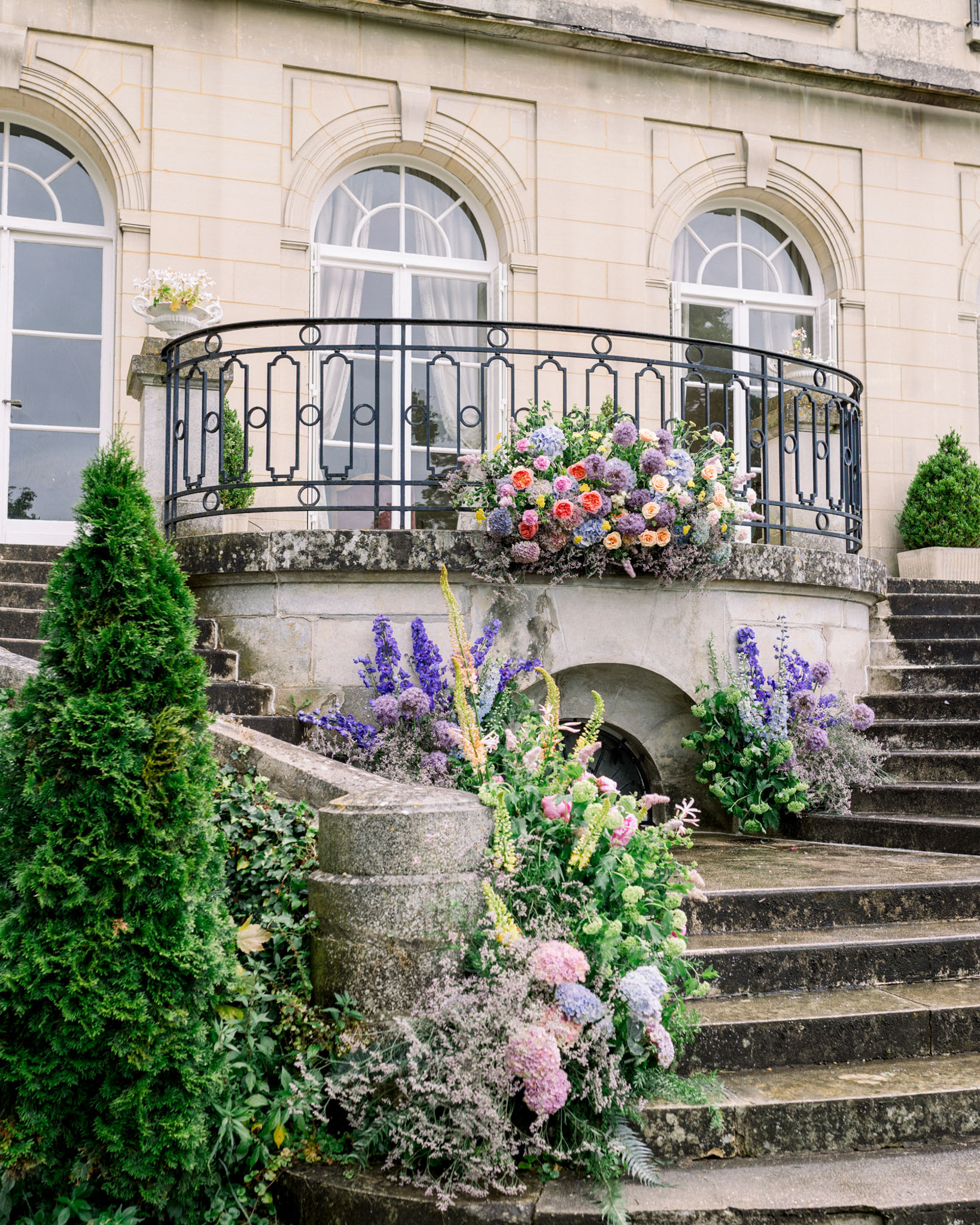 Classical stone building facade with arched windows and wrought-iron balcony decorated with coral, pink, and lavender wedding florals