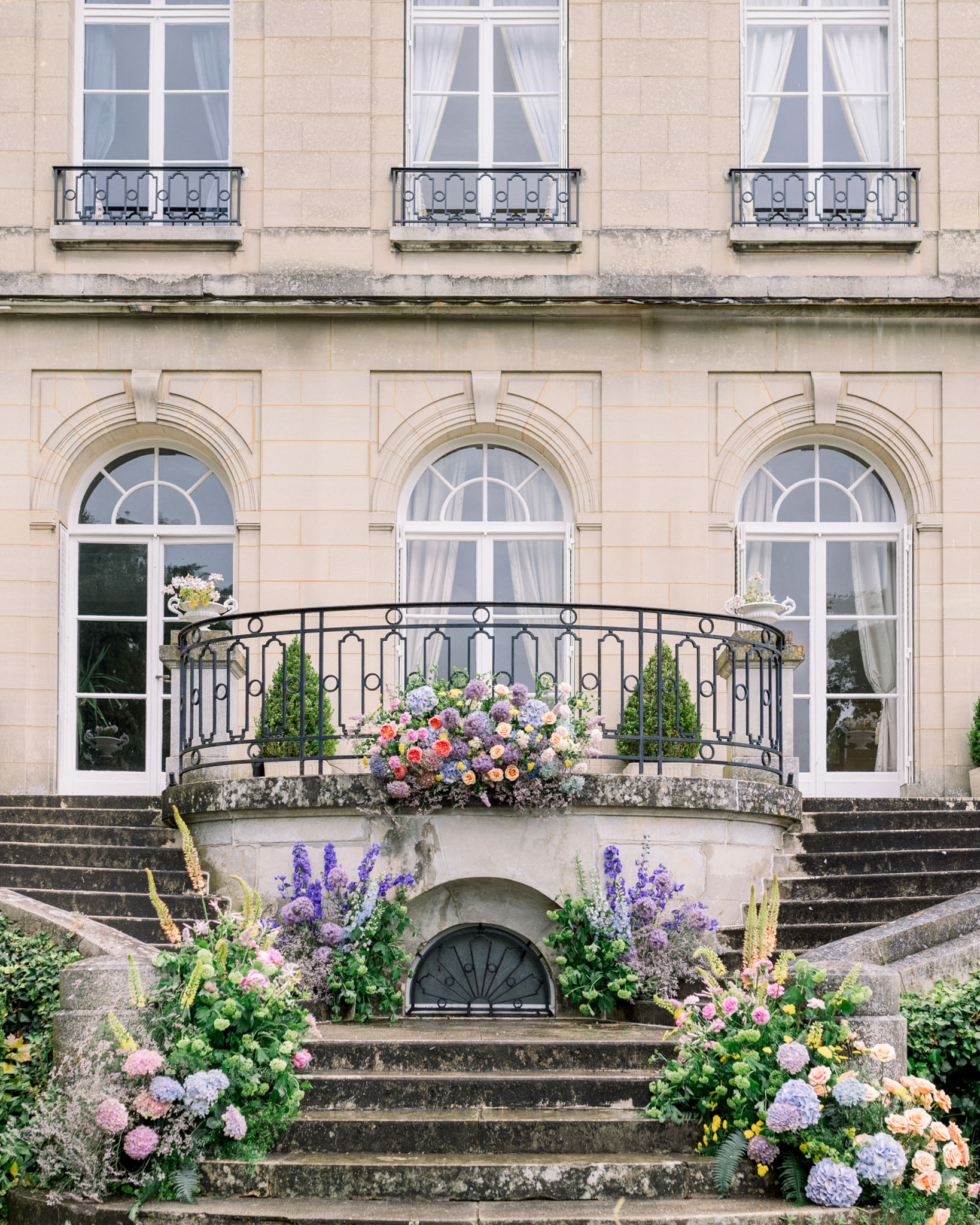 Neoclassical stone façade with curved wrought-iron balcony decorated with pink hydrangeas and blue delphiniums