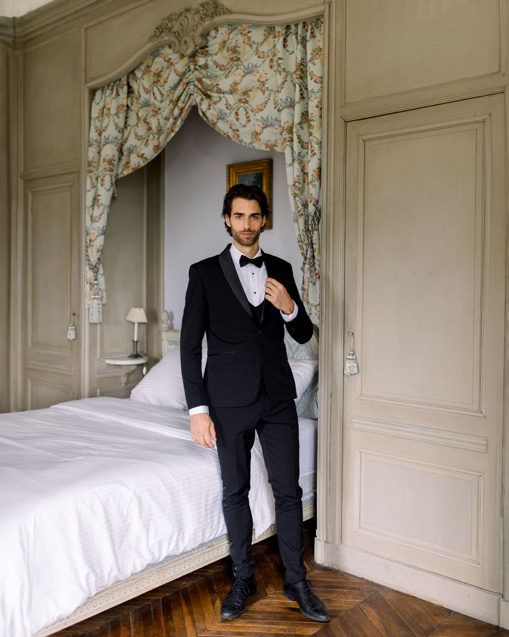 Groom in black tuxedo and bow tie adjusting jacket in doorway of classical bedroom with sage-green paneled walls