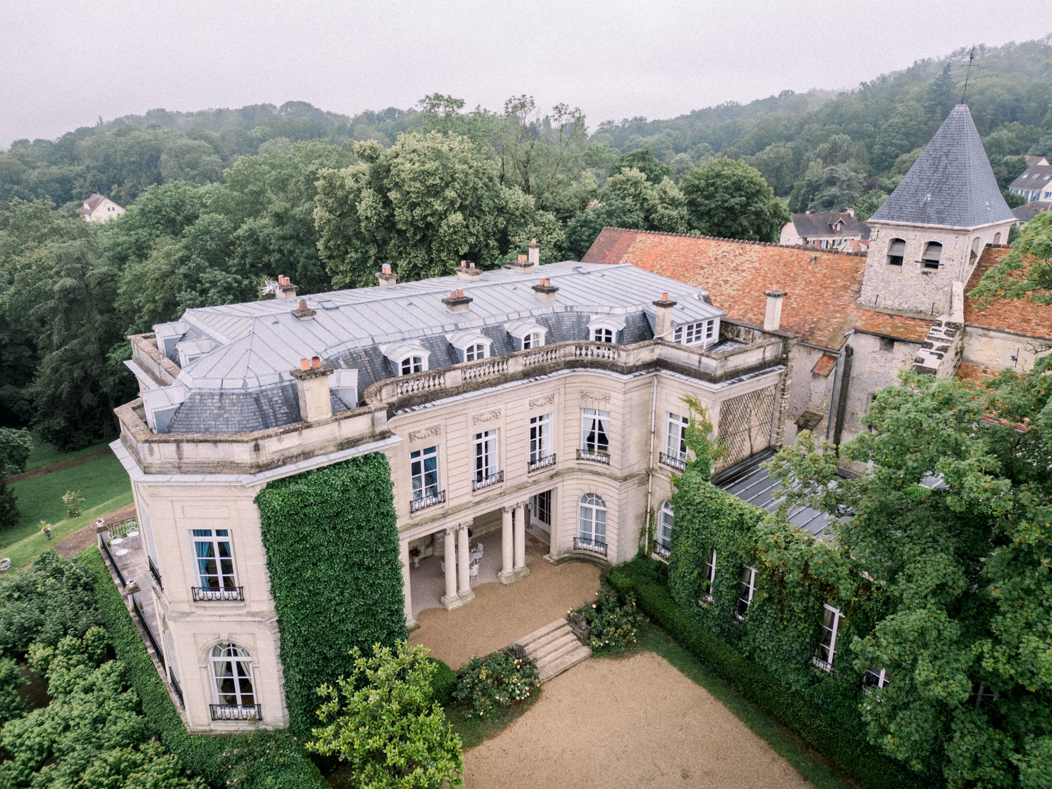Aerial view of classical French château with mansard roofs and round tower set within forested valley