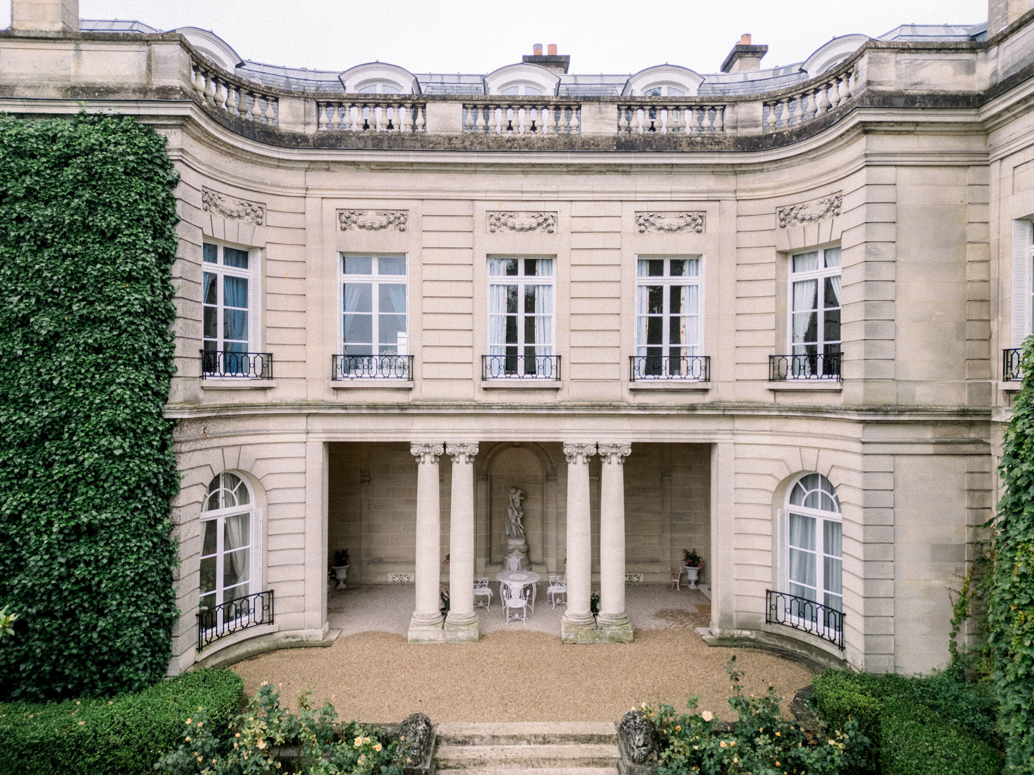 A wide exterior shot of a French neoclassical mansion or hôtel particulier, photographed from an elevated angle looking toward the curved rear façade. The building features cream-colored limestone construction with ornate carved stone detailing above the windows, wrought-iron Juliet balconies on the upper floor, and a ground-level covered colonnade with Ionic columns framing a central arched niche housing a white classical statue. A small white wrought-iron garden table and chairs are positioned beneath the colonnade. The forecourt is finished in gravel, and the foreground includes formally clipped low hedging and a rose bush with pale yellow blooms. No people are visible in the image. Potential venue feature image.