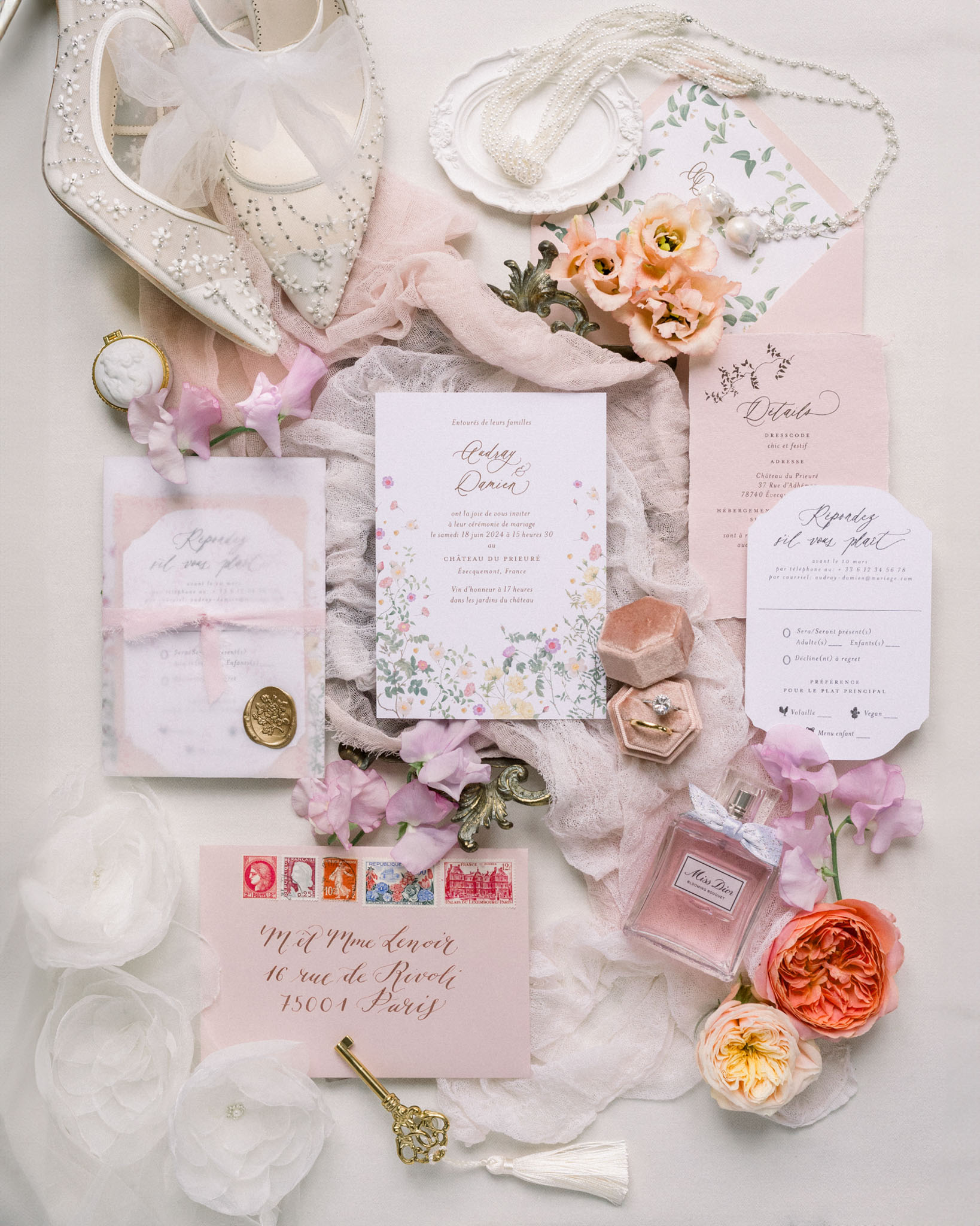 Flat lay of wedding stationery, ivory beaded shoes, pink clutch, perfume, and coral roses on neutral background